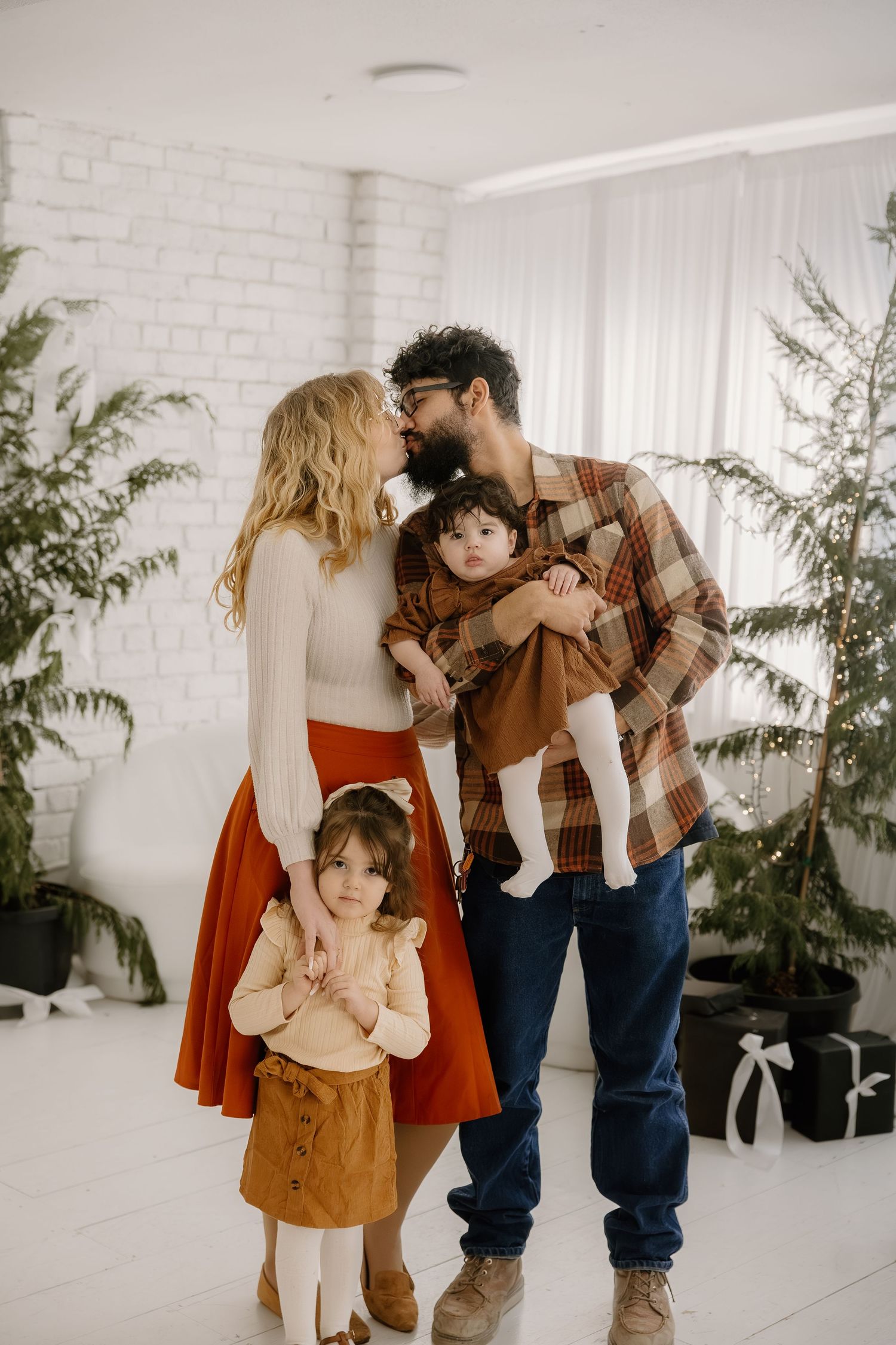 Young family sharing a kiss while holding two small children in a bright white decorated room.