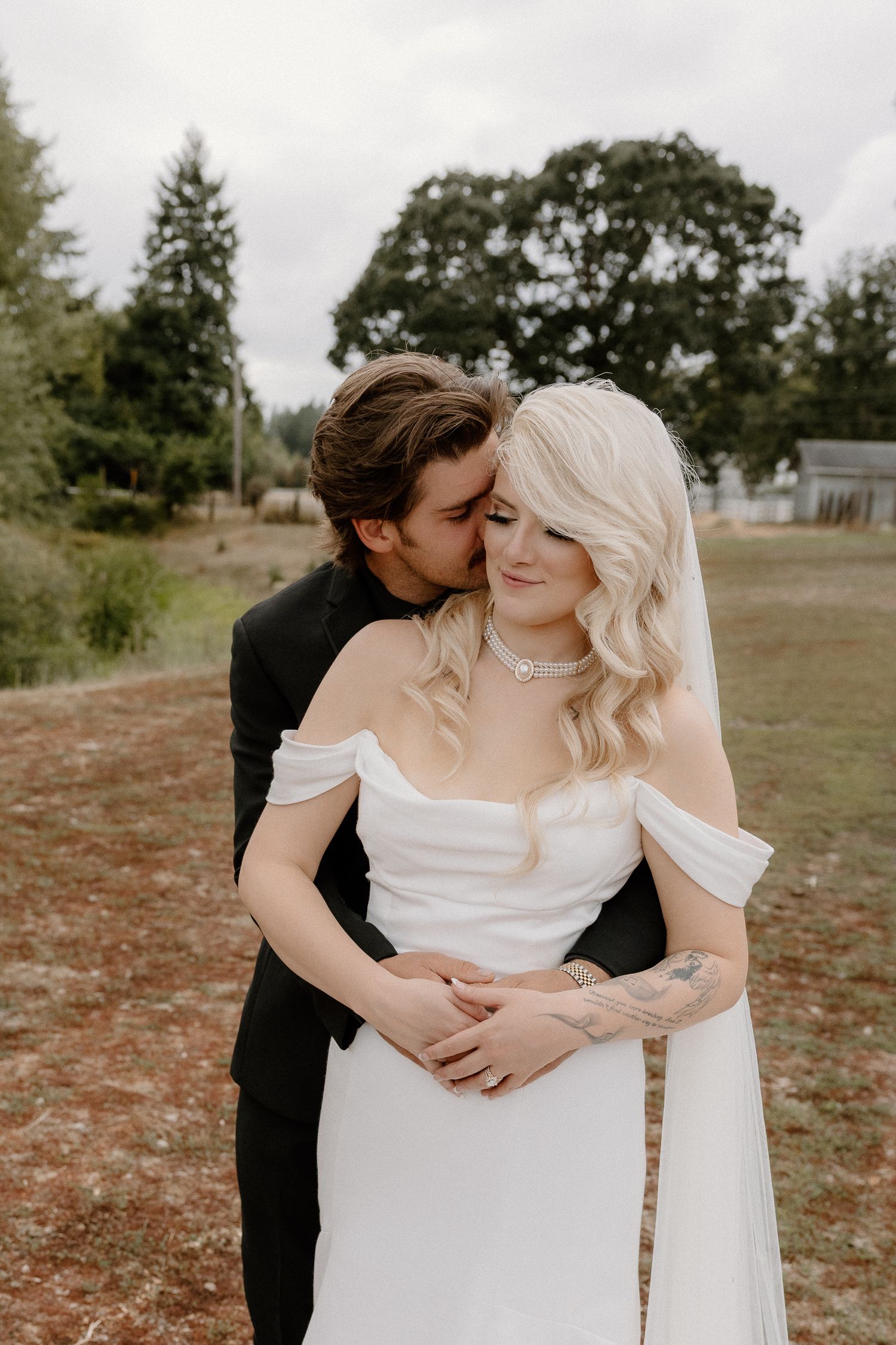 Groom embracing bride from behind and kissing her cheek at outdoor rustic wedding venue.