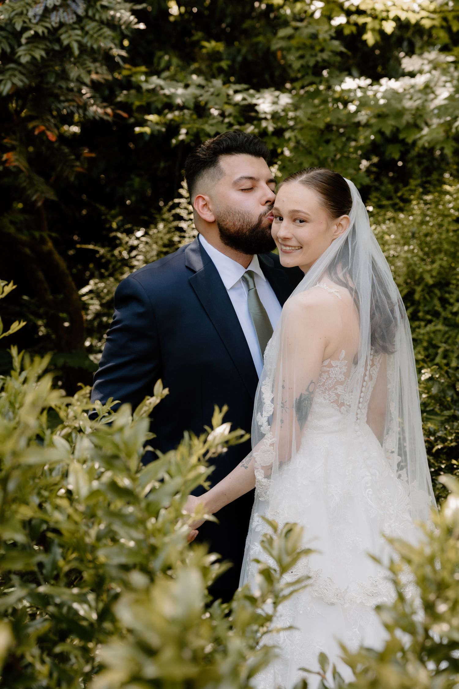 Groom in navy suit kisses bride's cheek as she smiles in lush garden setting.