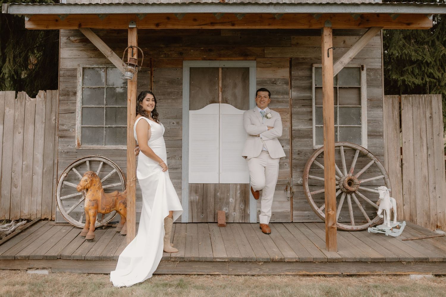 Bride and groom posing on rustic wooden porch with wagon wheels and dog on wedding day.