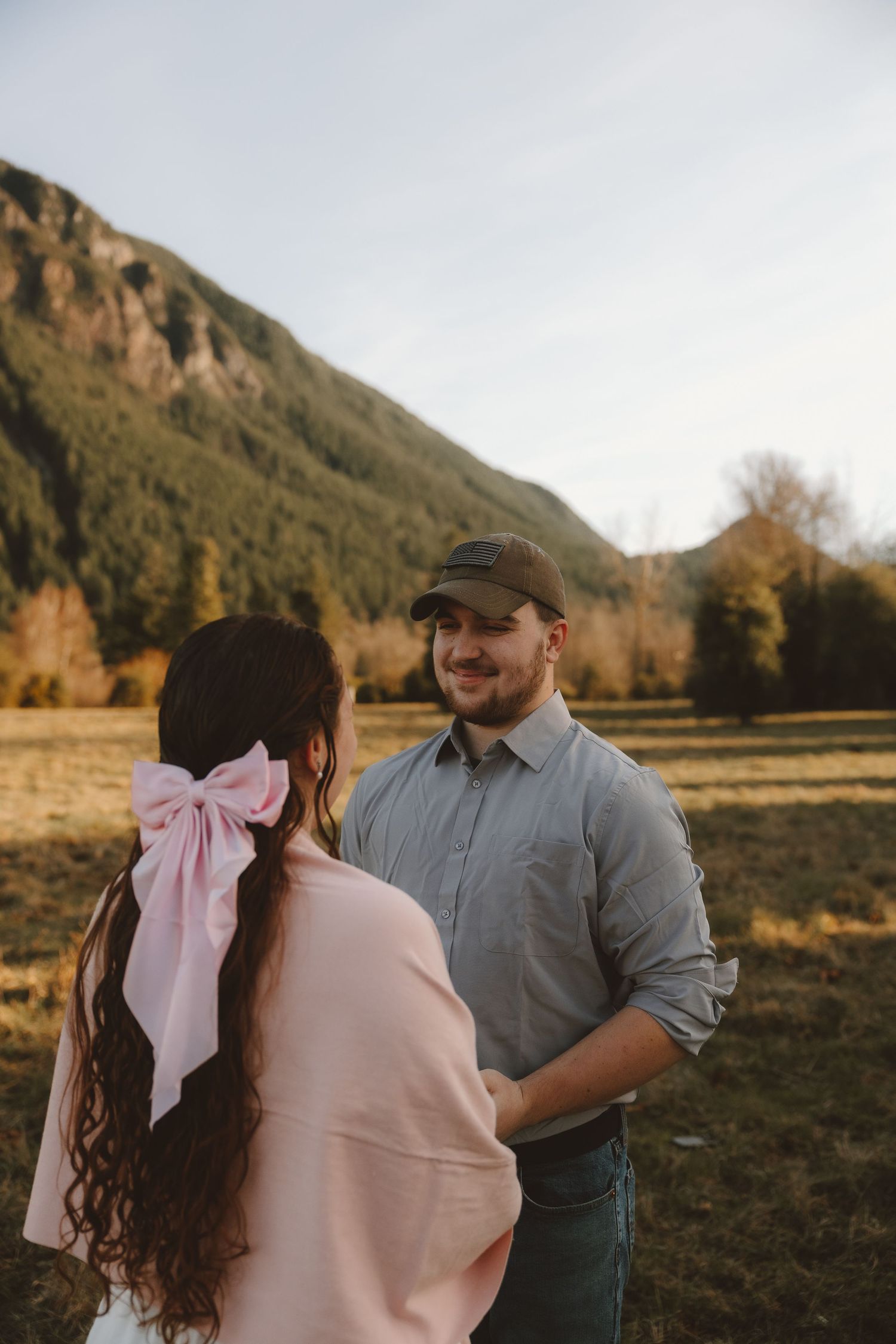 Couple sharing a romantic moment in a mountain meadow, woman with pink bow in hair facing man in gray shirt and cap.