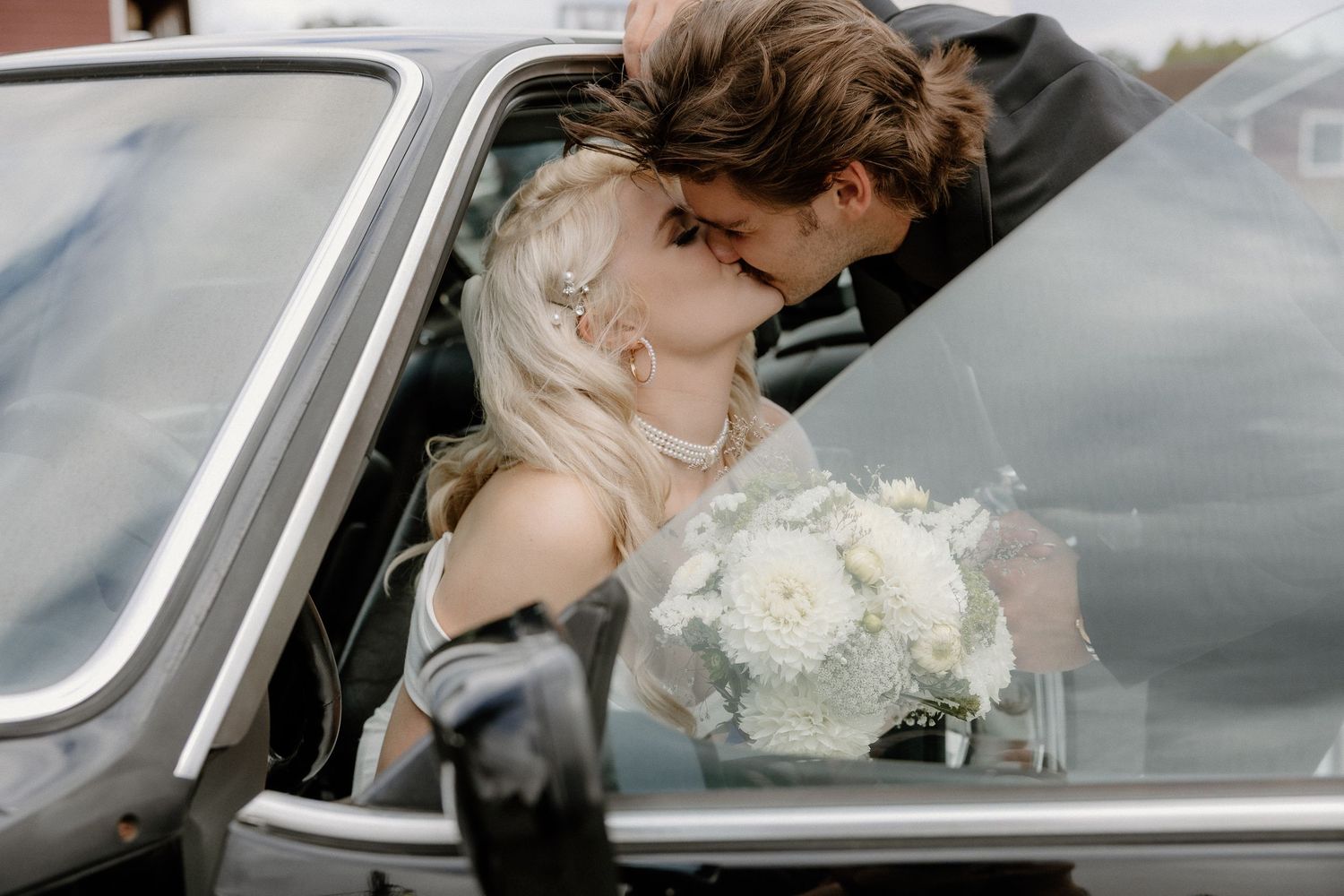 Newlyweds kissing inside vintage car with bride holding white floral bouquet on wedding day.