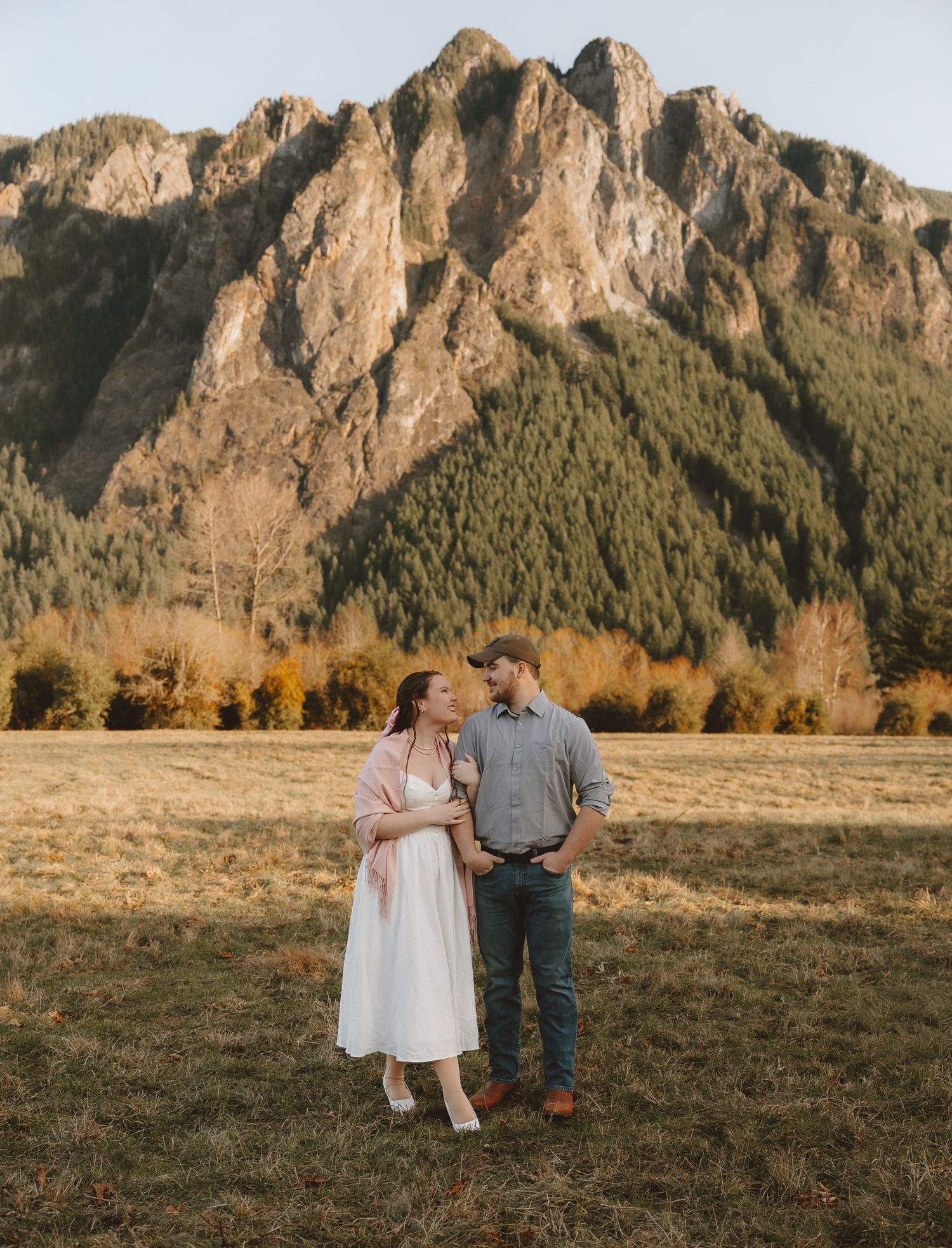 Couple embracing in a golden meadow with a dramatic rocky mountain and autumn trees in the background.