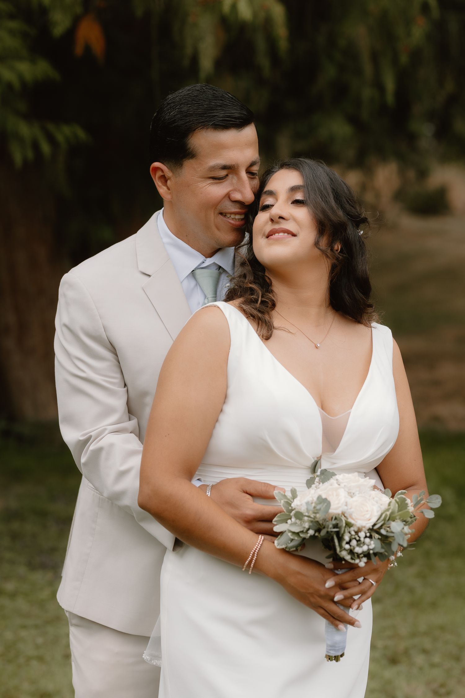 Groom in a beige suit embraces his bride in a white dress, holding a white bouquet outdoors.