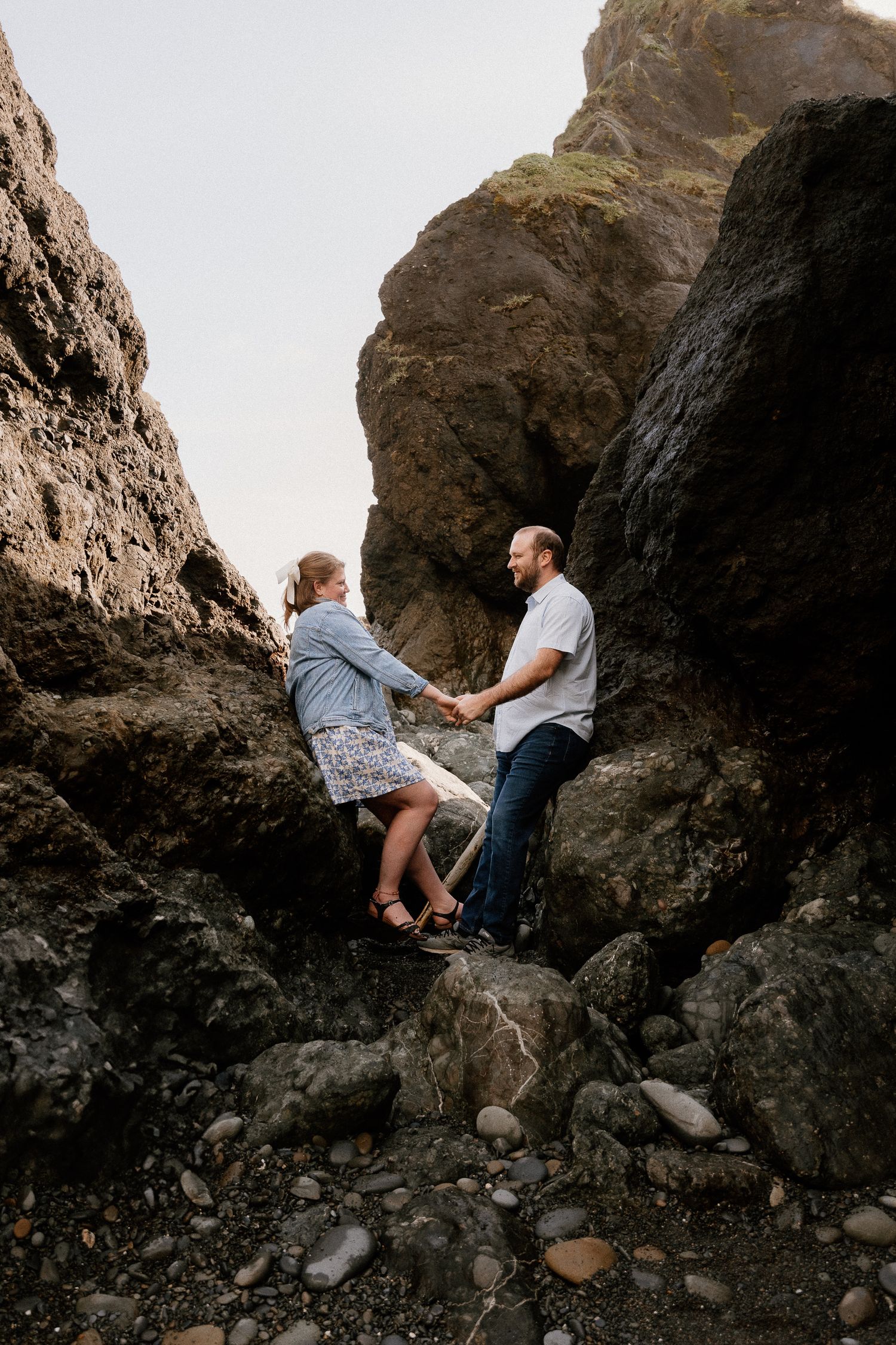 Couple holding hands on rocky coastal cliffs, wearing casual attire, surrounded by large boulders and pebble beach below.