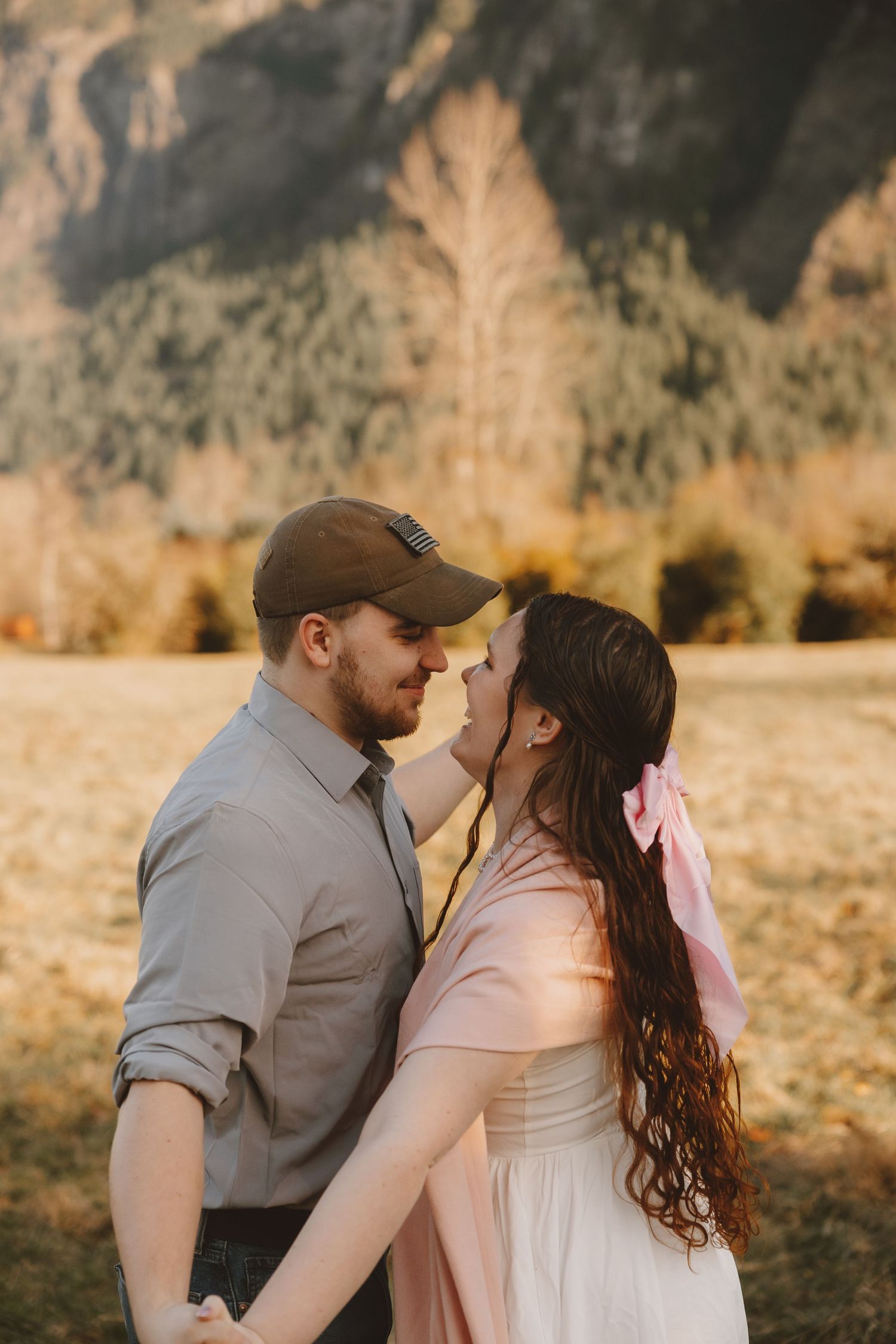 Couple sharing a romantic moment outdoors with mountain backdrop, woman in pink dress with bow, man in gray shirt and cap.