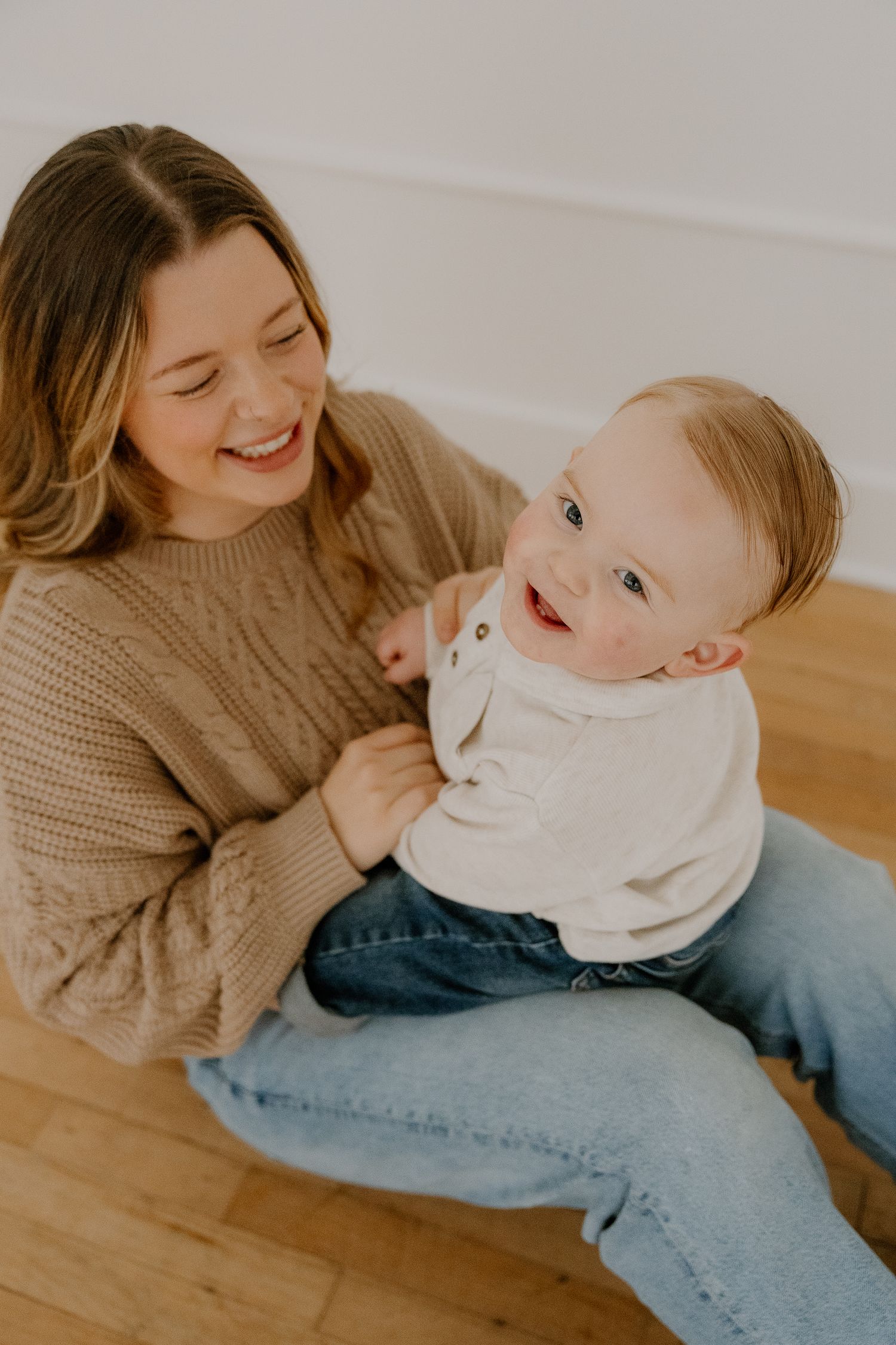 Joyful mother in cable knit sweater sits on floor laughing with smiling baby on her lap.