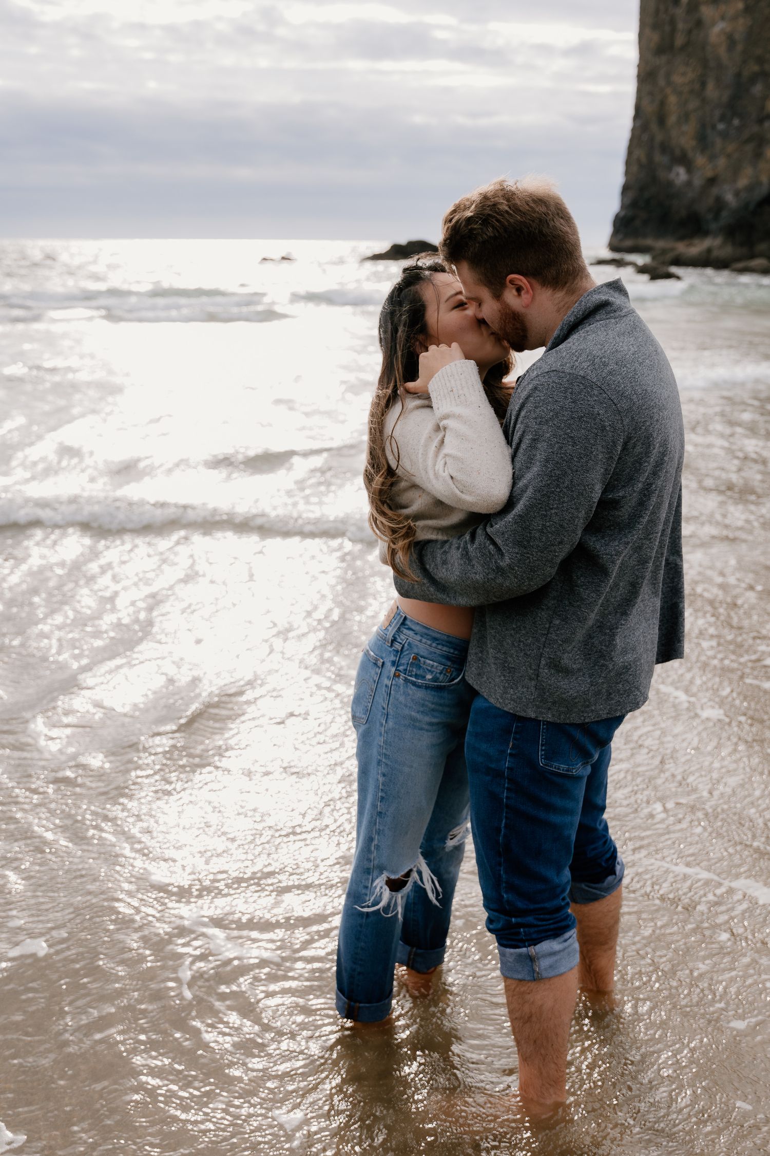 Couple embracing and kissing on a wet beach shore with ocean waves and rocky cliffs in the background.