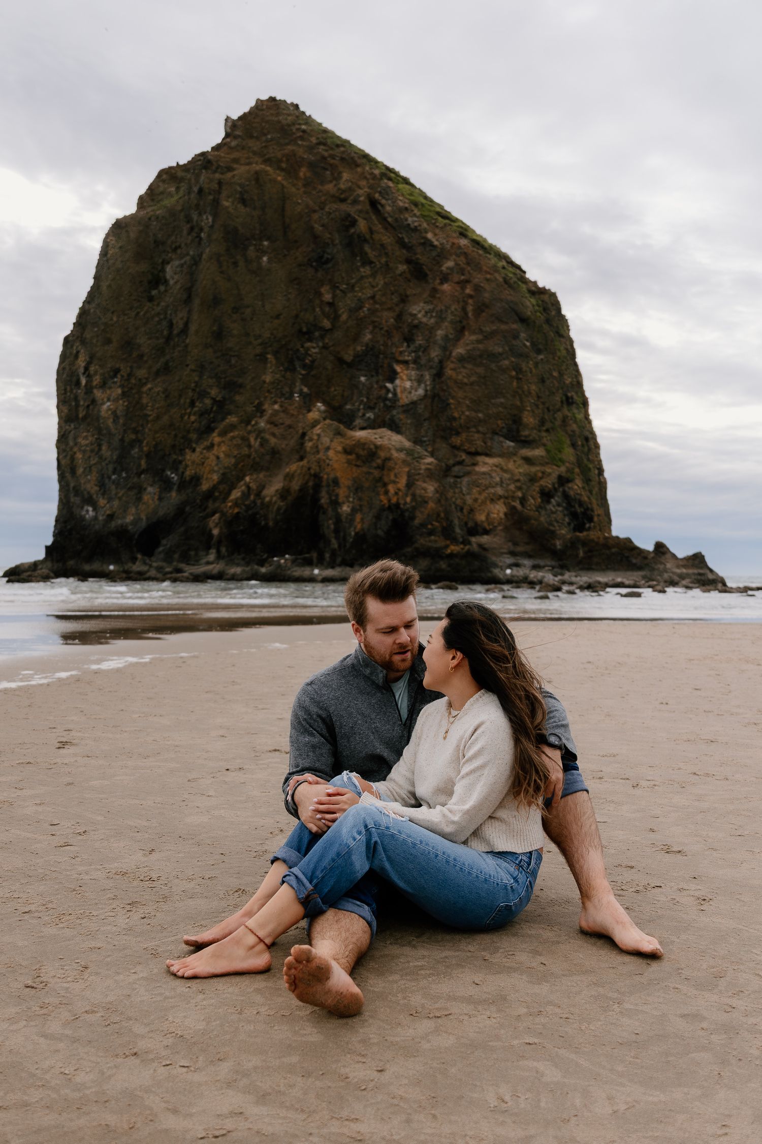 Couple sitting barefoot on sandy beach in front of iconic Haystack Rock at Cannon Beach, Oregon on a cloudy day.
