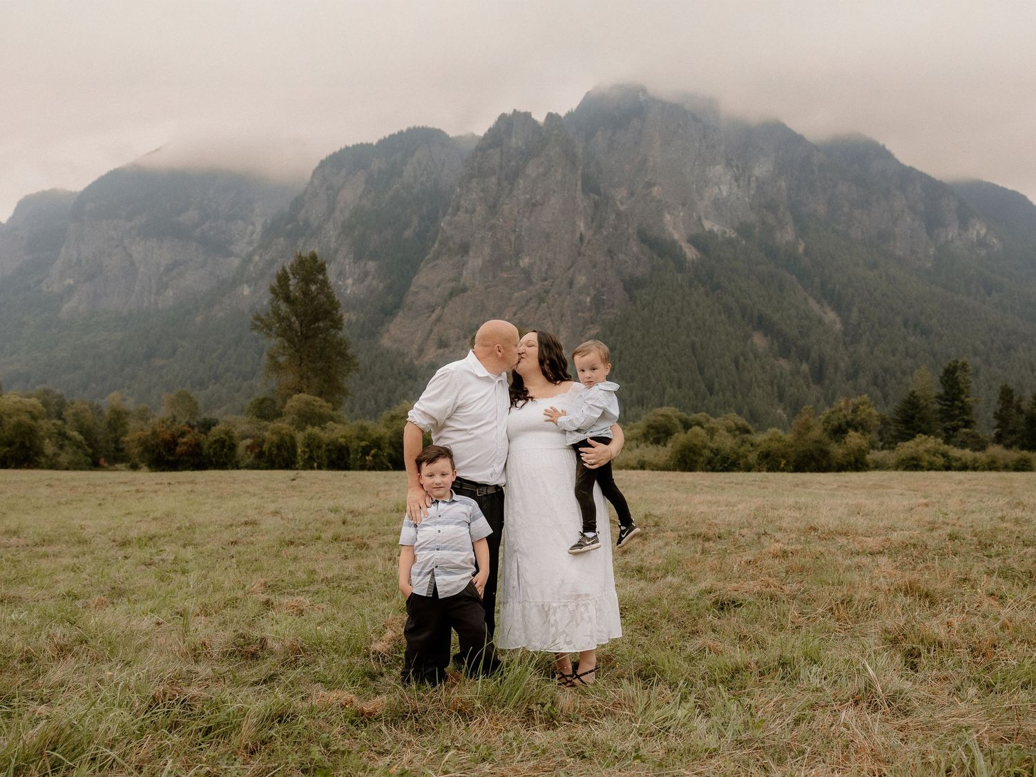 Family of four embraces in open field with dramatic misty mountain backdrop in Pacific Northwest.