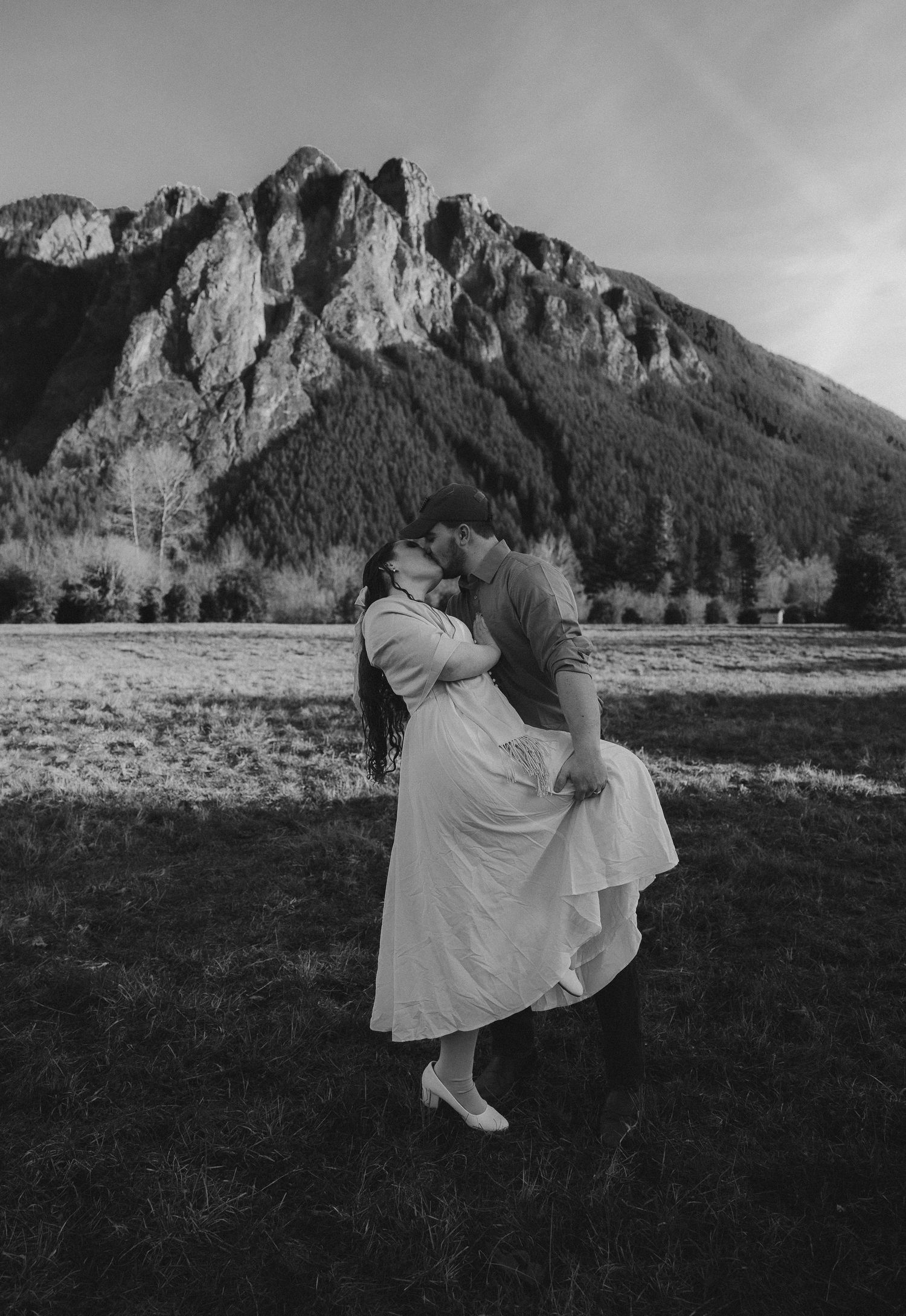 Black and white romantic photo of couple kissing in field with mountain scenery behind them.