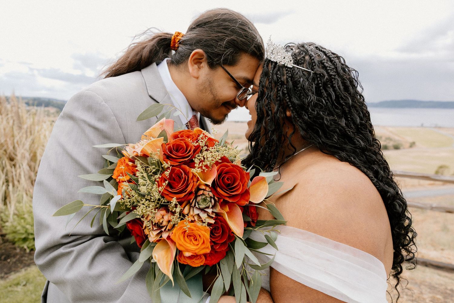 Couple touching foreheads romantically while holding vibrant orange and red rose wedding bouquet outdoors.