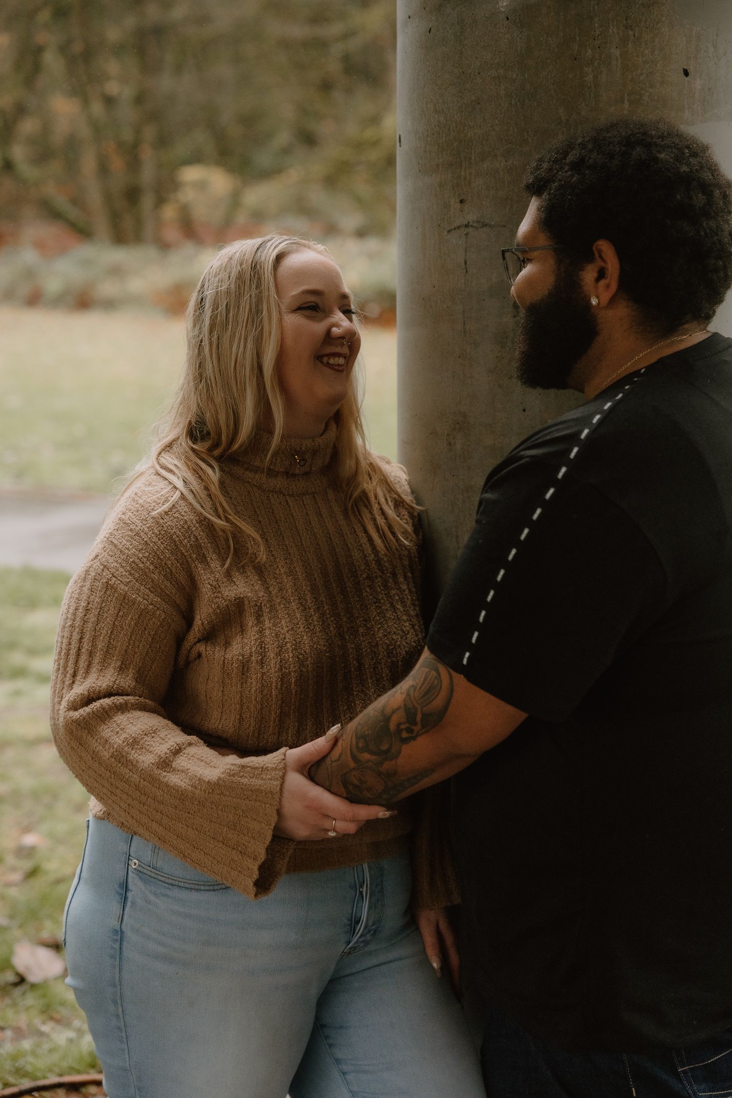 A couple holding hands and smiling at each other beside a tree in an outdoor park setting.