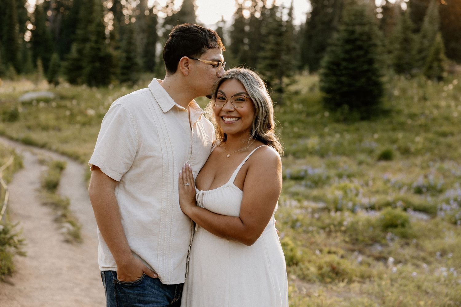 A man kissing a smiling woman on the head in a sunlit alpine meadow with pine trees.