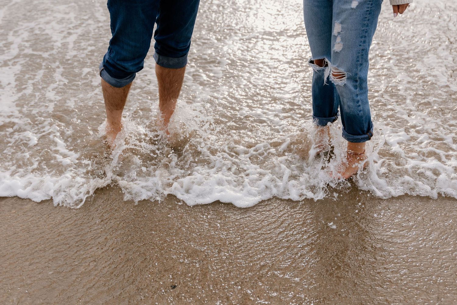 Two people in rolled-up jeans walking barefoot in ocean waves on a sandy beach.