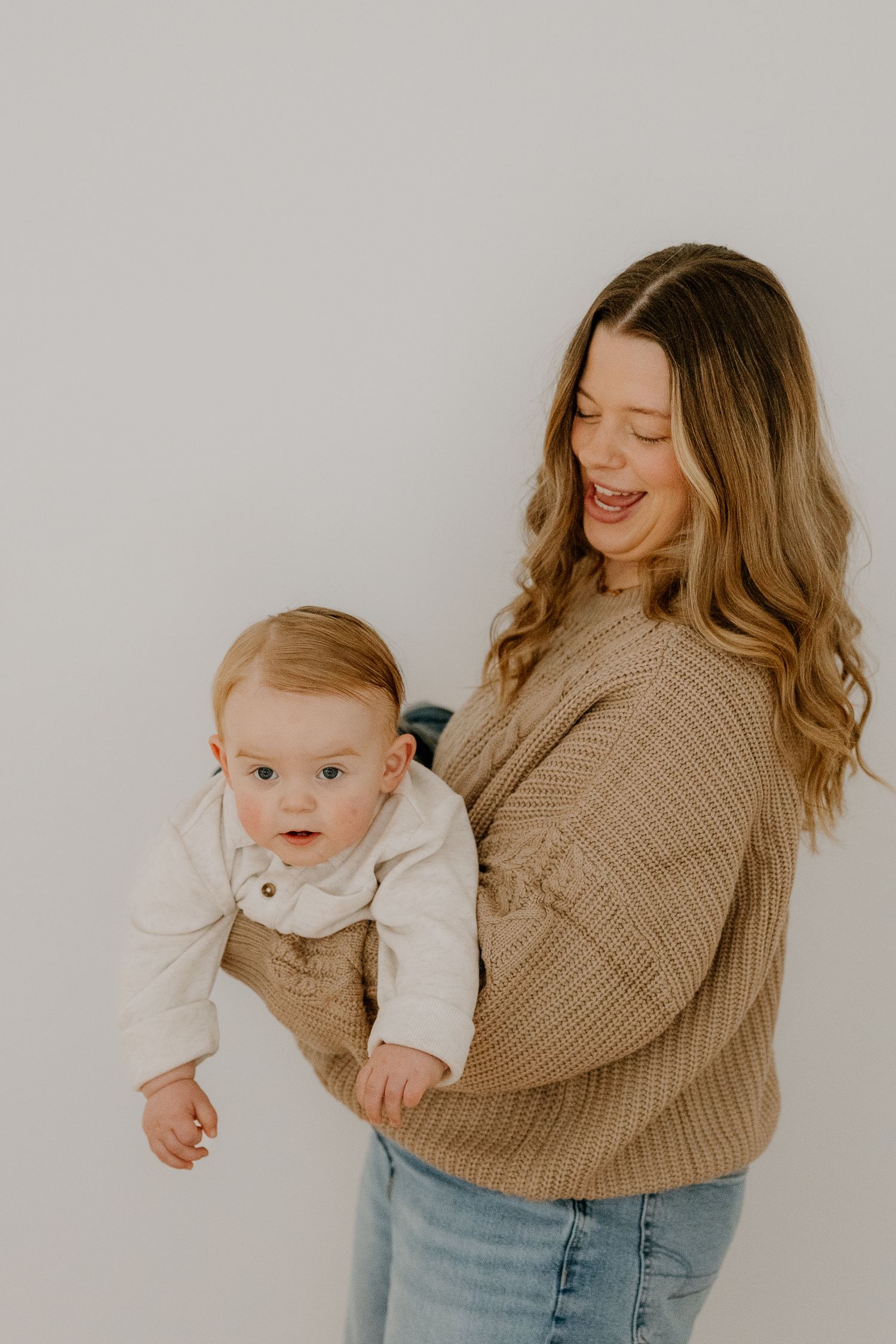 Cheerful mother in beige sweater playfully holds laughing baby outward against white background.