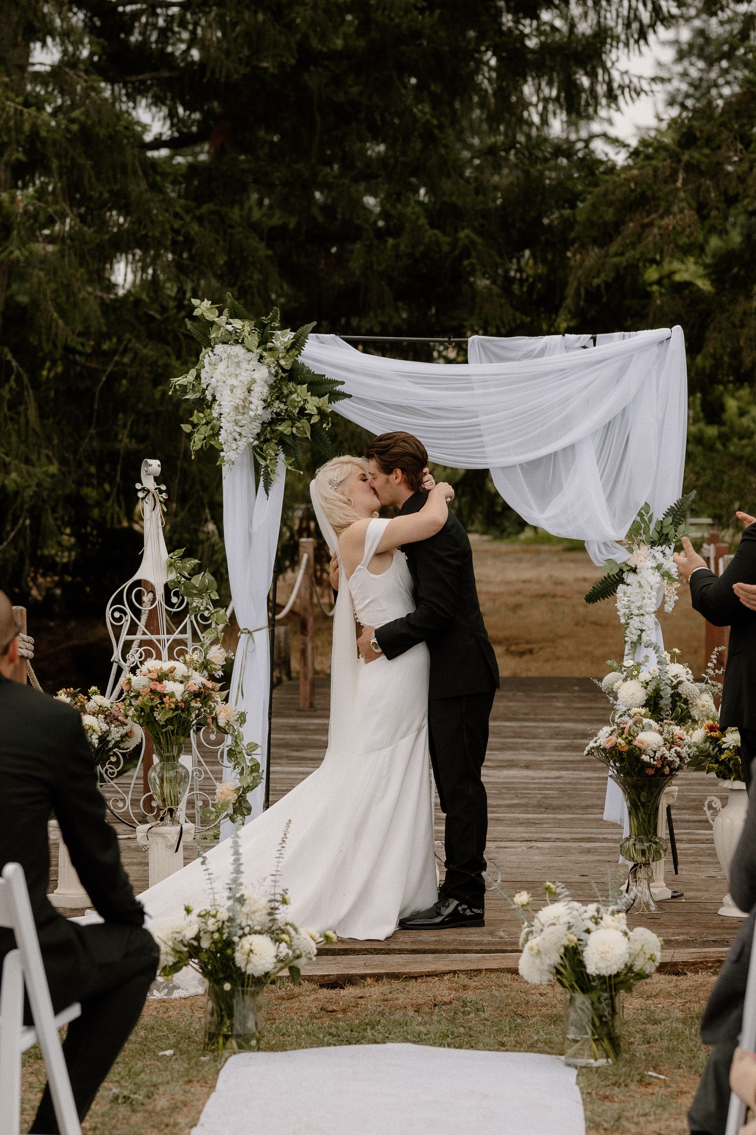 A bride and groom share their first kiss under a white draped arch at an outdoor woodland wedding.