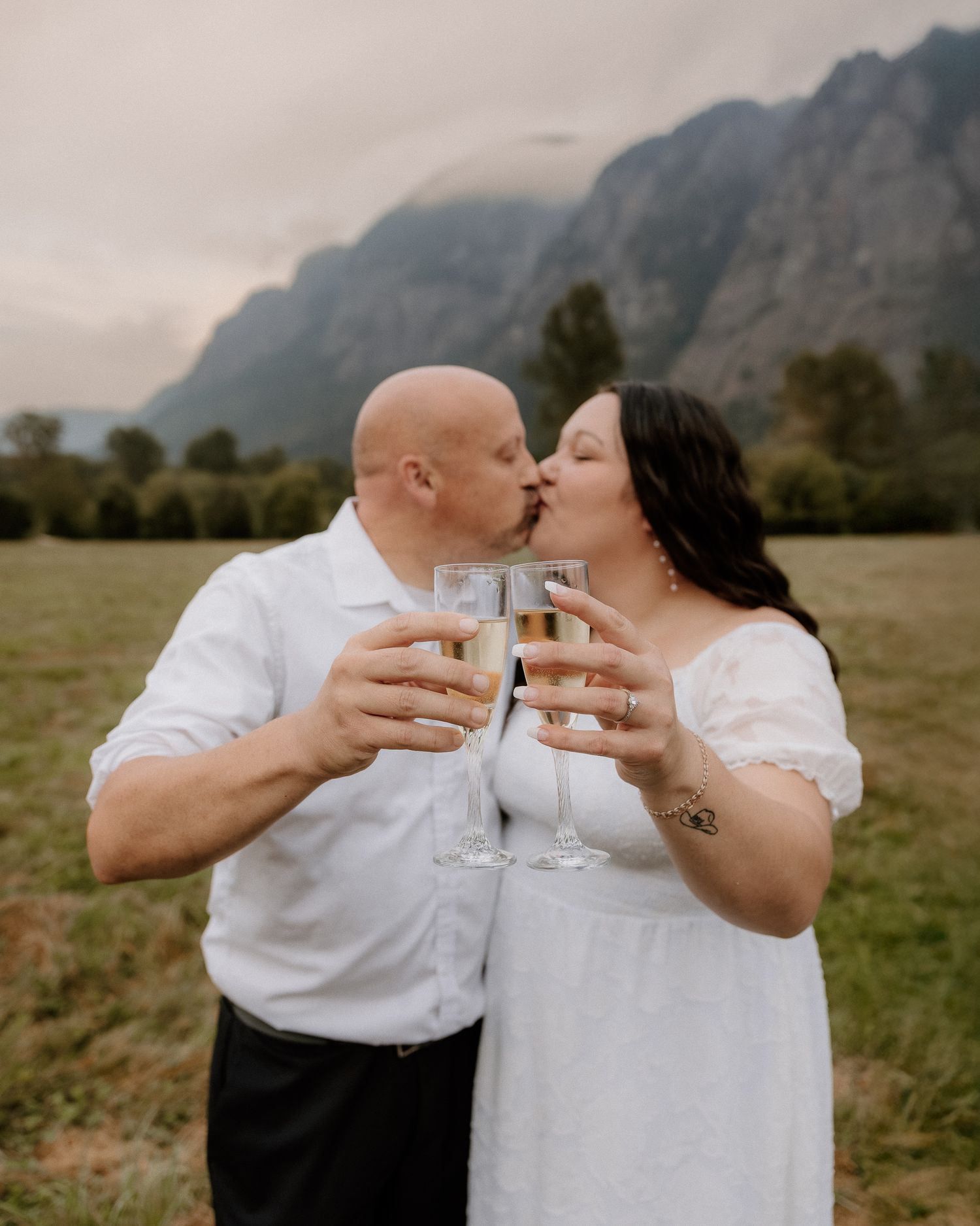 Couple kisses while toasting with champagne glasses in a field with dramatic mountain scenery behind them.