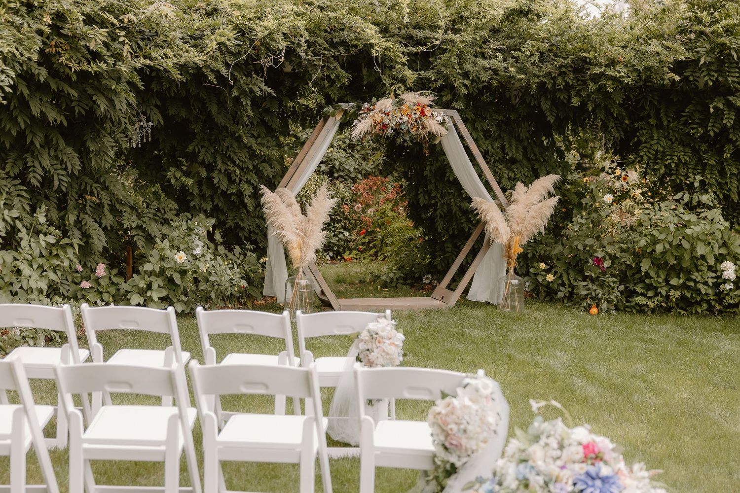 Outdoor wedding ceremony setup with hexagonal arch decorated with pampas grass and white chairs on green lawn.