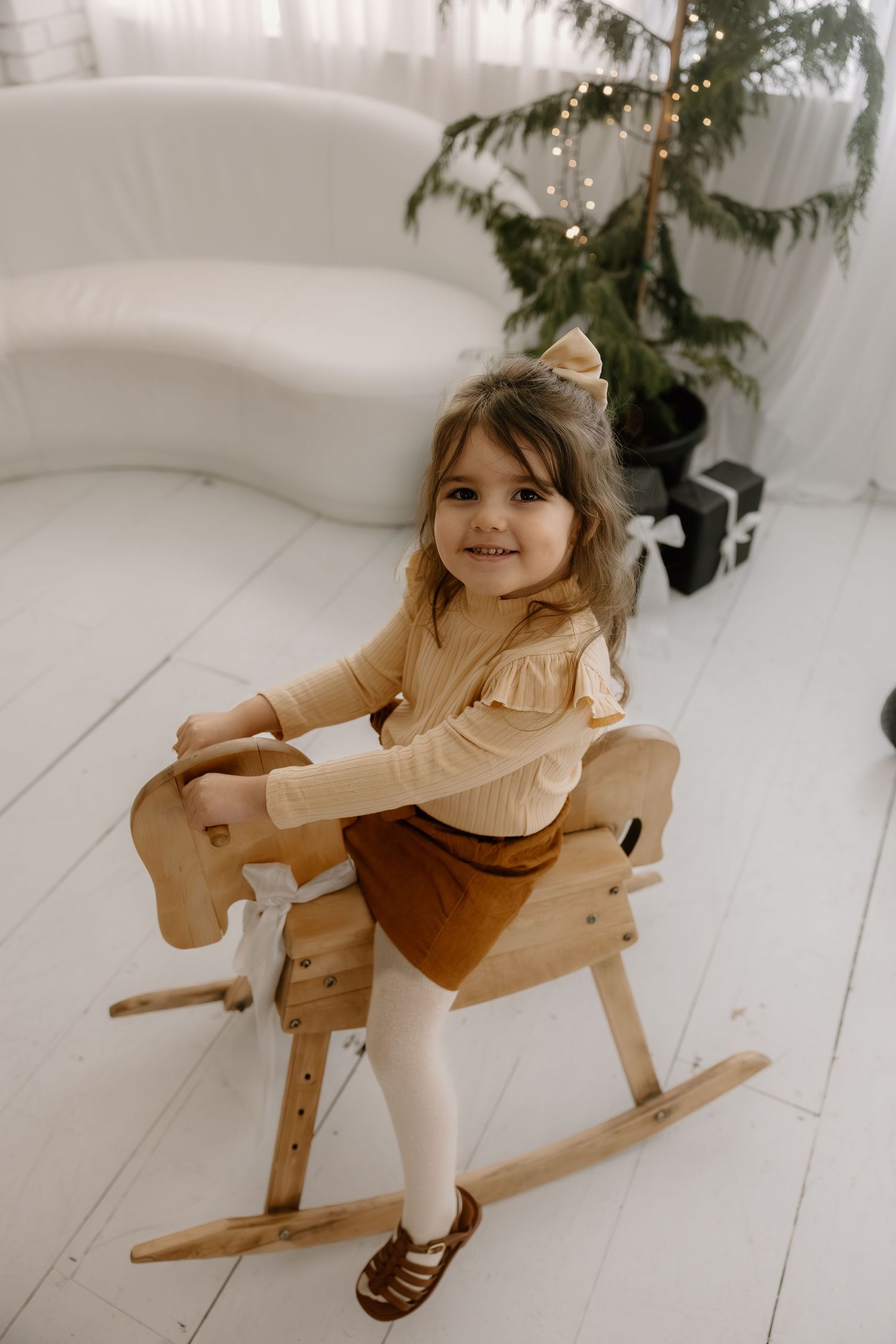 Smiling little girl in brown outfit sitting on a wooden rocking horse near a Christmas tree.