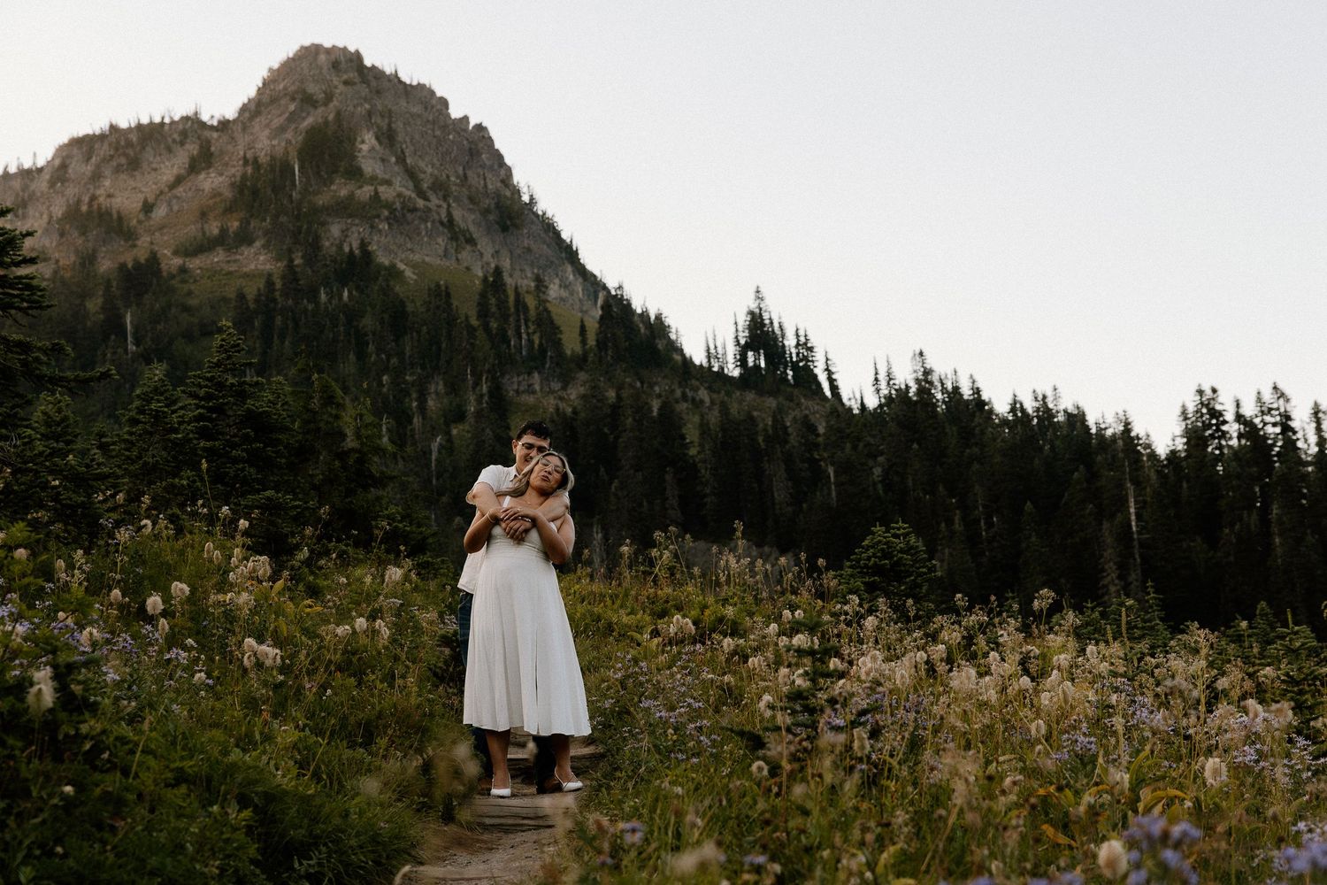 A couple embracing on a rocky trail surrounded by wildflowers and a dramatic mountain peak.