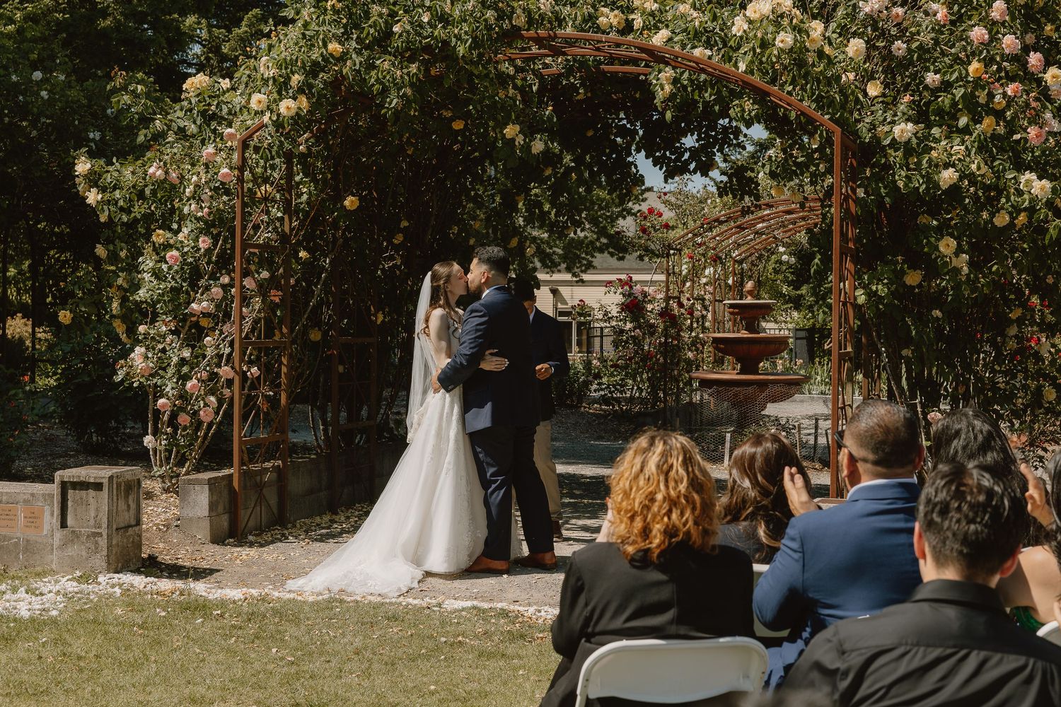 A couple shares their first kiss under a floral arch during an outdoor garden wedding ceremony.