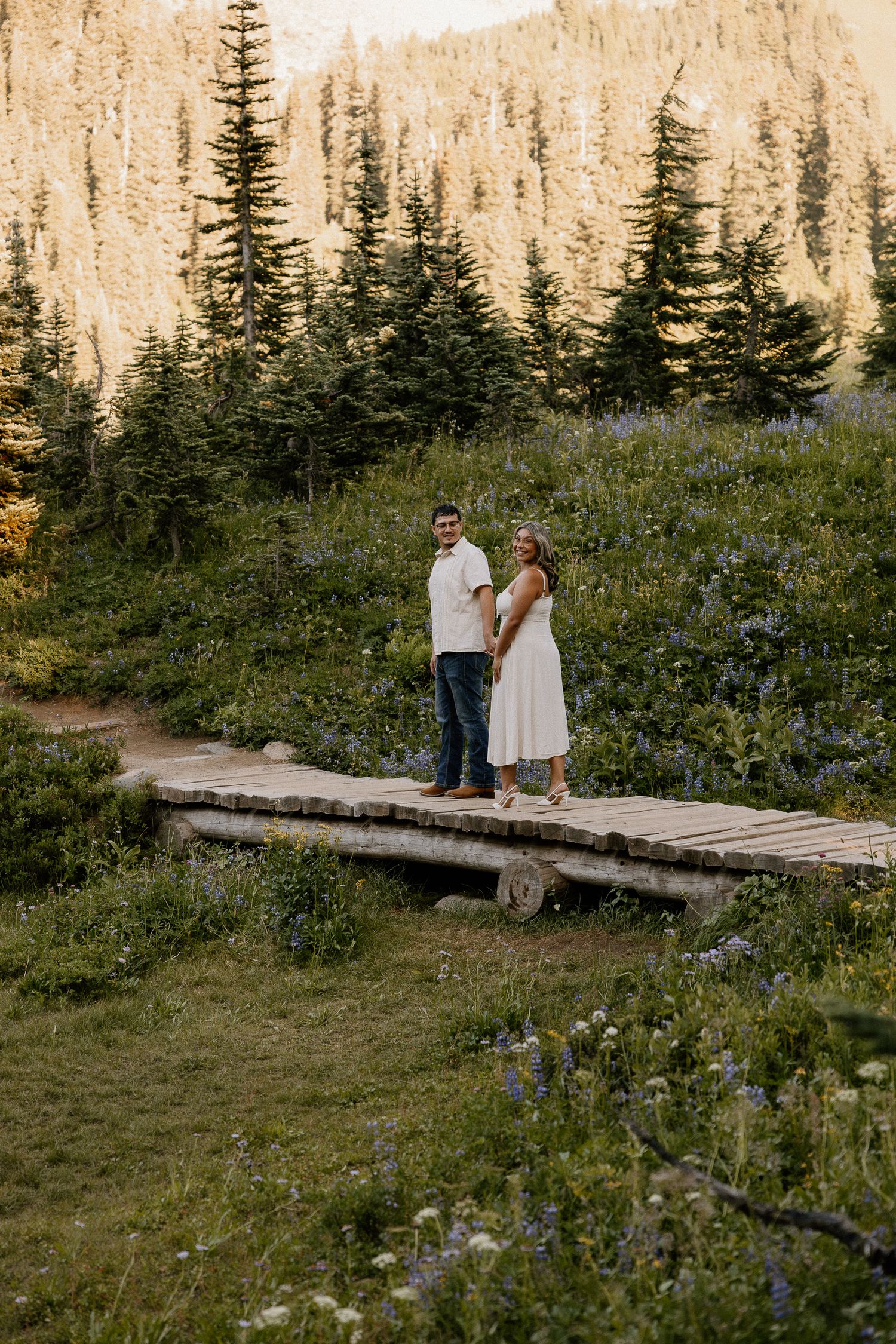 A couple holds hands on a wooden bridge surrounded by wildflowers and pine trees at golden hour.