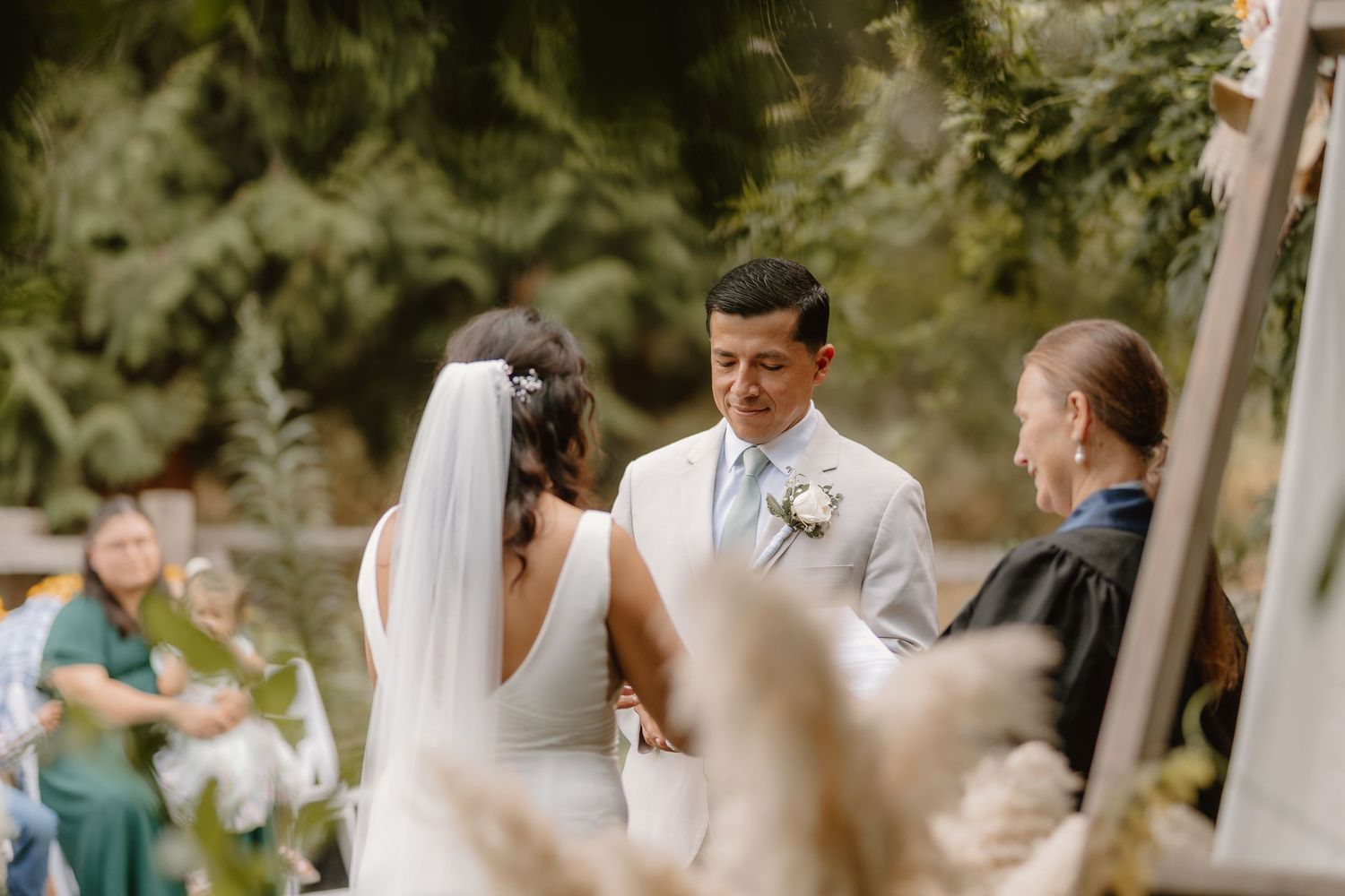 Bride and groom exchange vows outdoors surrounded by lush greenery and pampas grass decorations.