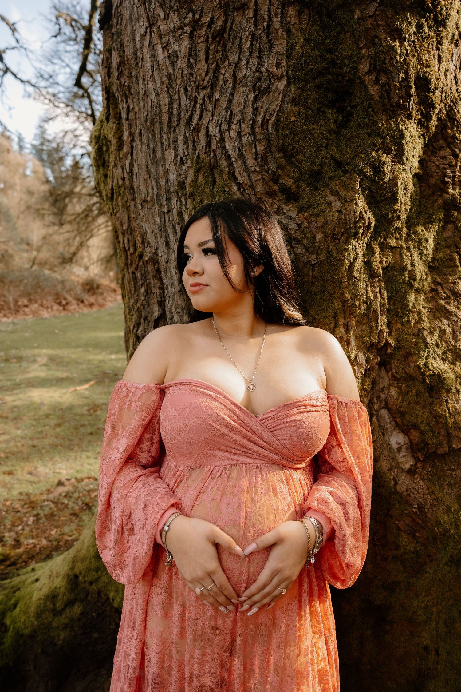 Pregnant woman in blush lace gown making heart shape over belly while leaning against large tree.