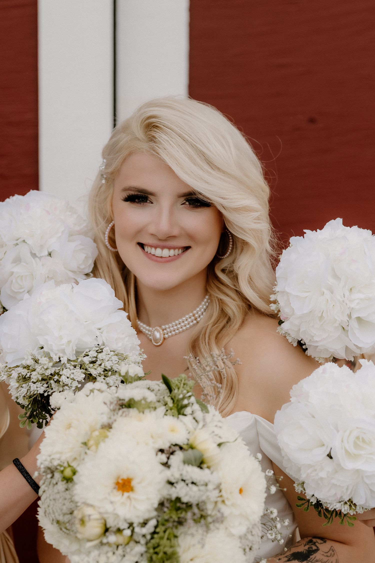 Smiling blonde bride wearing pearl choker necklace surrounded by white floral bouquets at her wedding.
