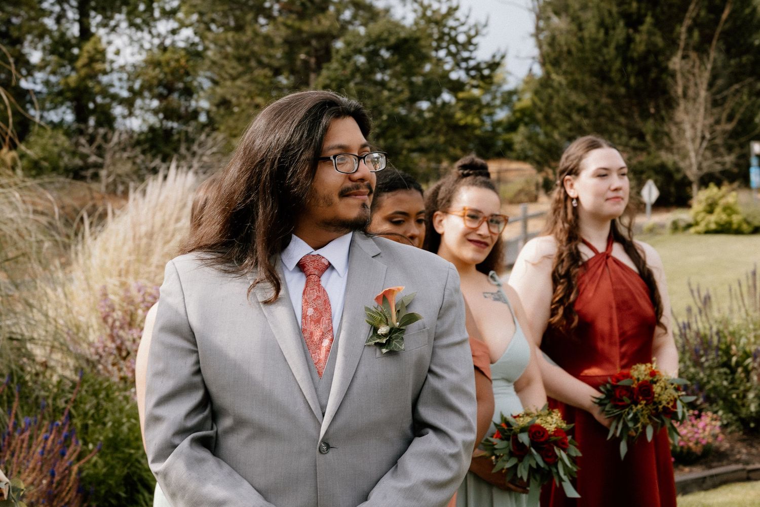 Groom in gray suit with orange boutonniere standing with wedding party in garden ceremony.