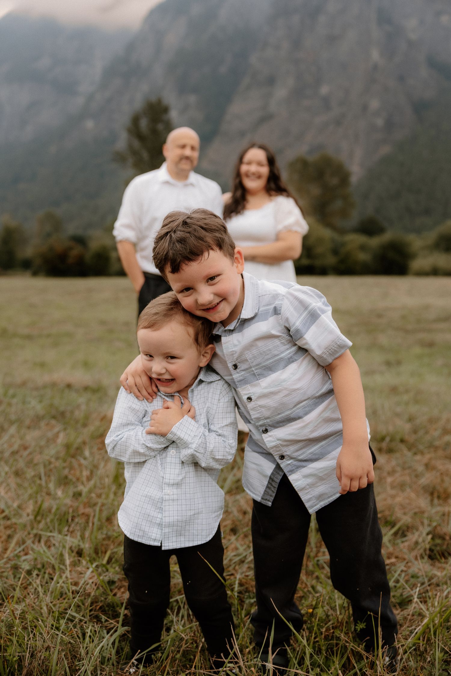 Two young brothers hugging in a mountain meadow with their smiling parents standing behind them.