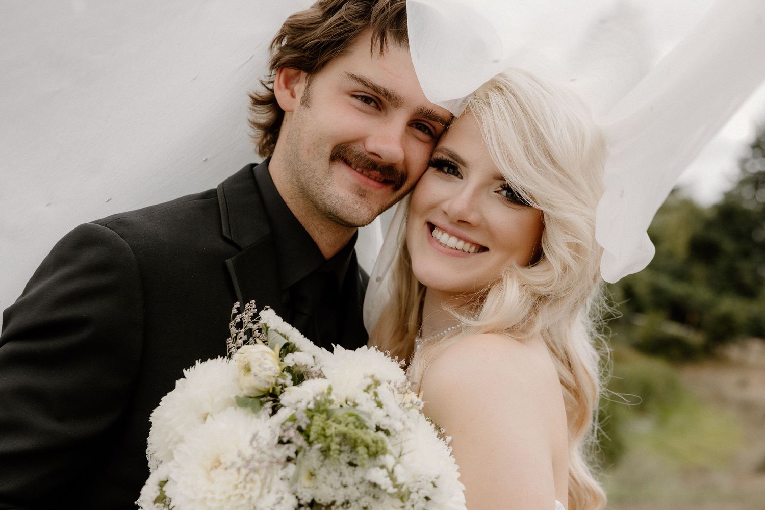 Smiling bride and groom pose closely together under a veil, with a white floral bouquet in a forest setting.
