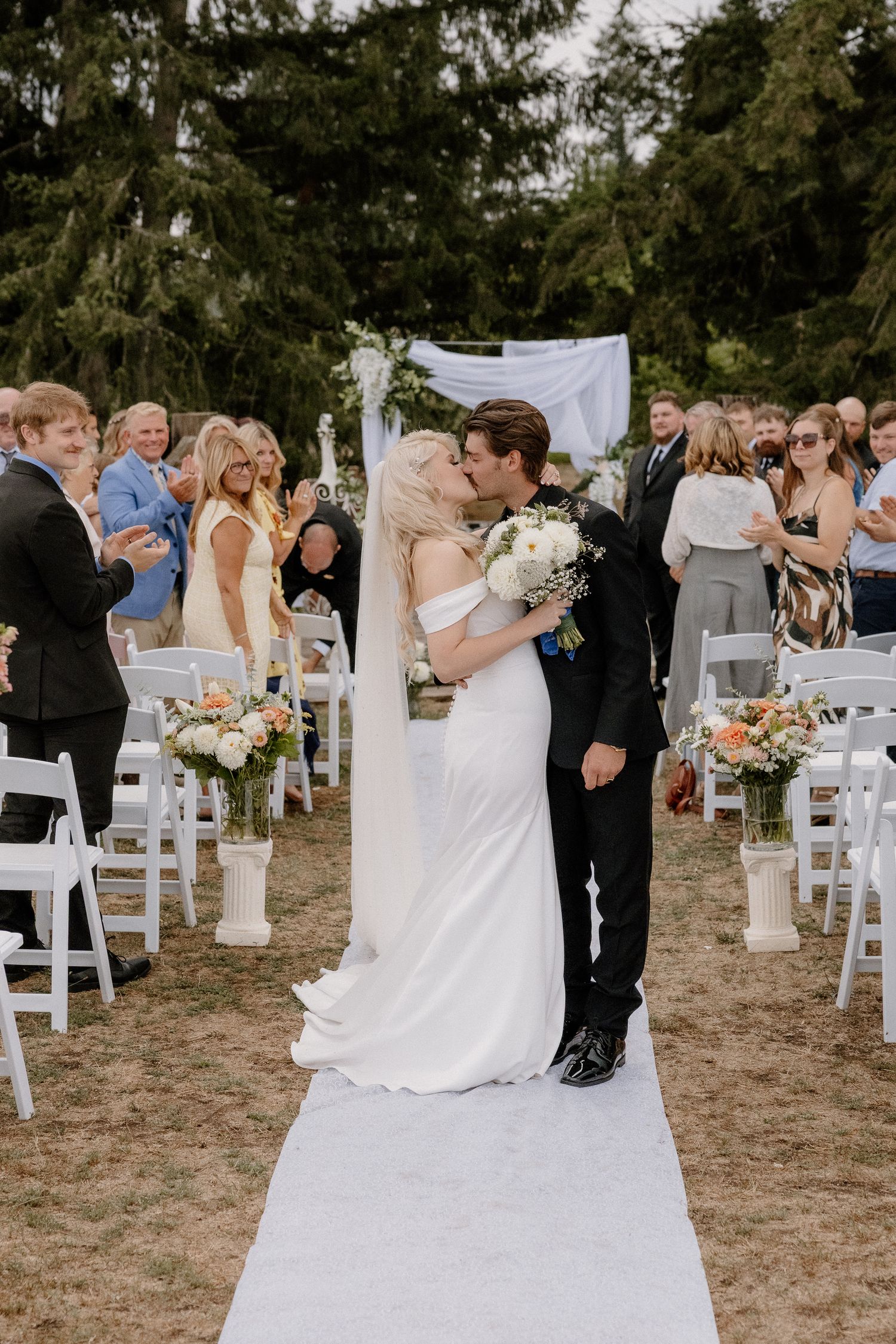 Newlyweds sharing first kiss at outdoor wedding ceremony aisle with guests applauding in background.