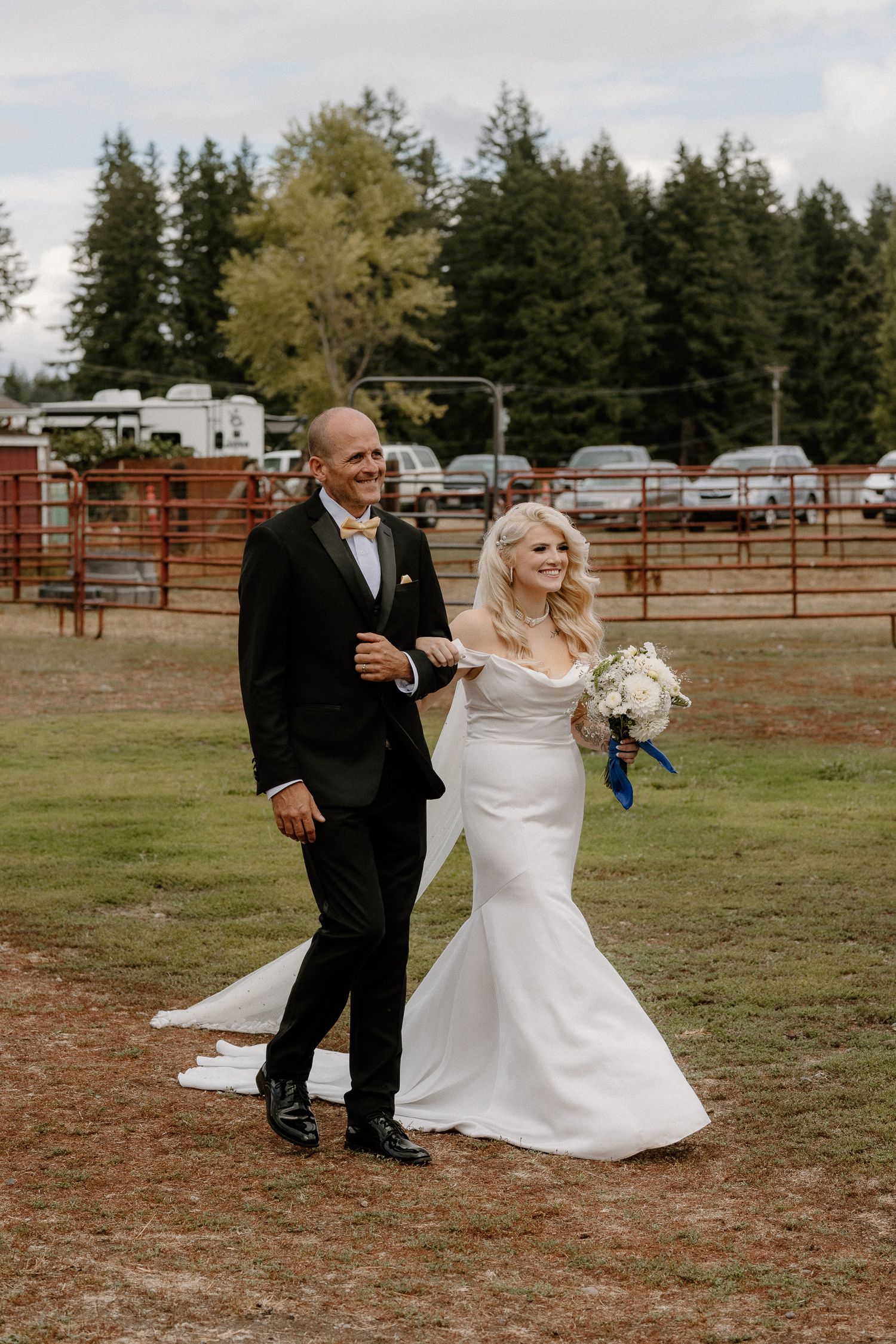 Bride and groom walking together outdoors at a rustic farm wedding venue, bride holding white bouquet with blue ribbon.