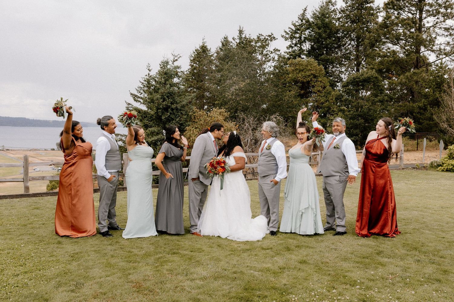 Wedding party celebrates outdoors with bridesmaids in sage and rust dresses as couple kisses center.