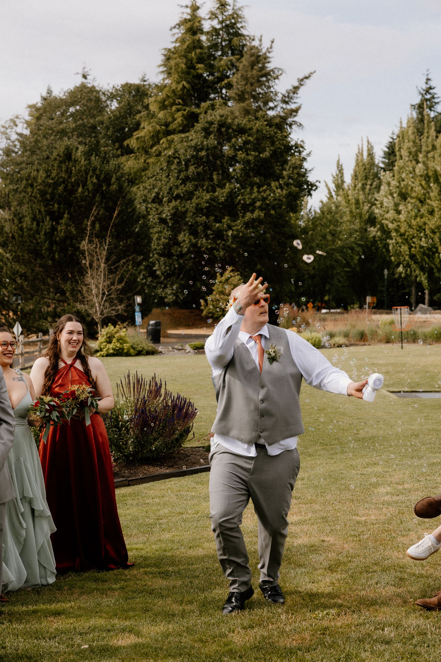 Best man in gray vest tosses bouquet on lawn as bridesmaids in red and sage dresses look on.