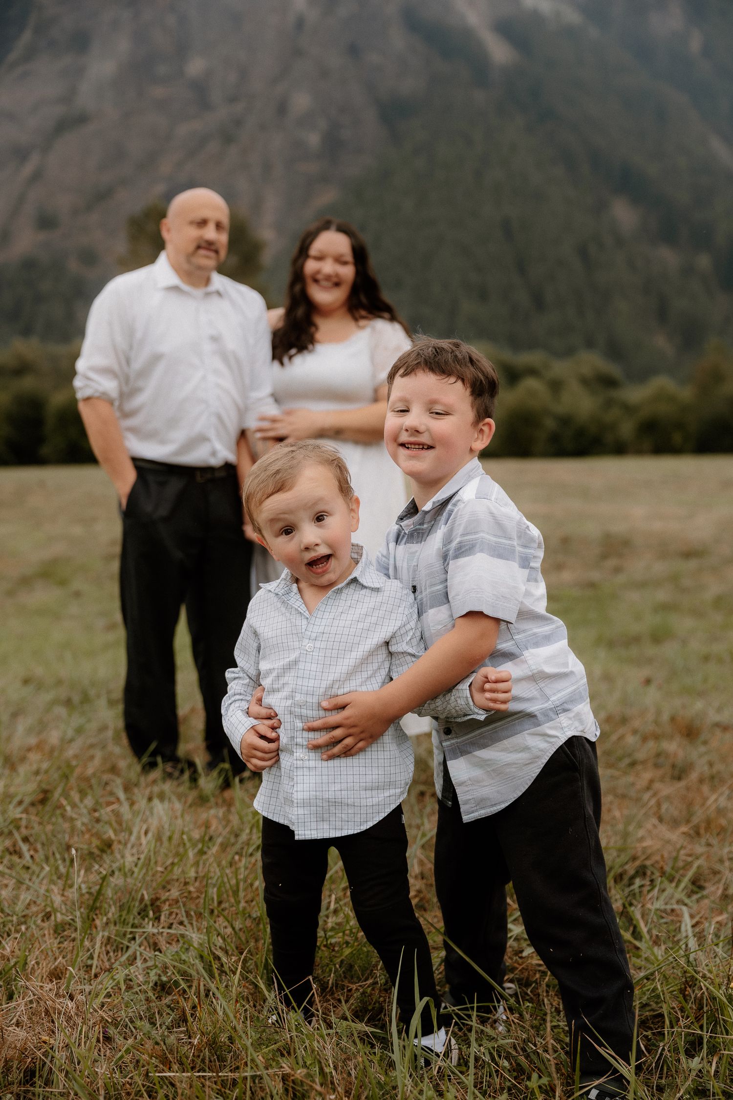 Two young boys laugh and play in a mountain meadow while their parents stand smiling behind them.