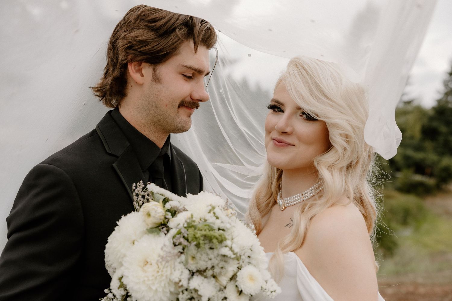 Bride and groom smiling at each other under flowing veil at outdoor wedding ceremony.