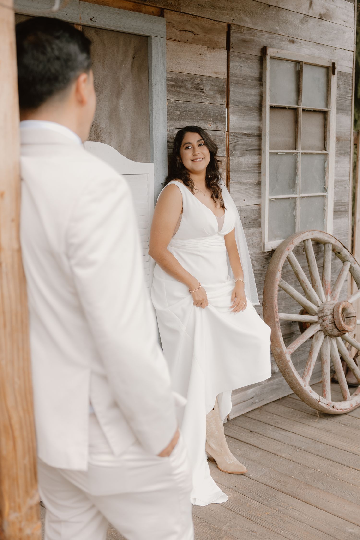 Smiling bride in white dress stands on rustic wooden porch with wagon wheel during first look.