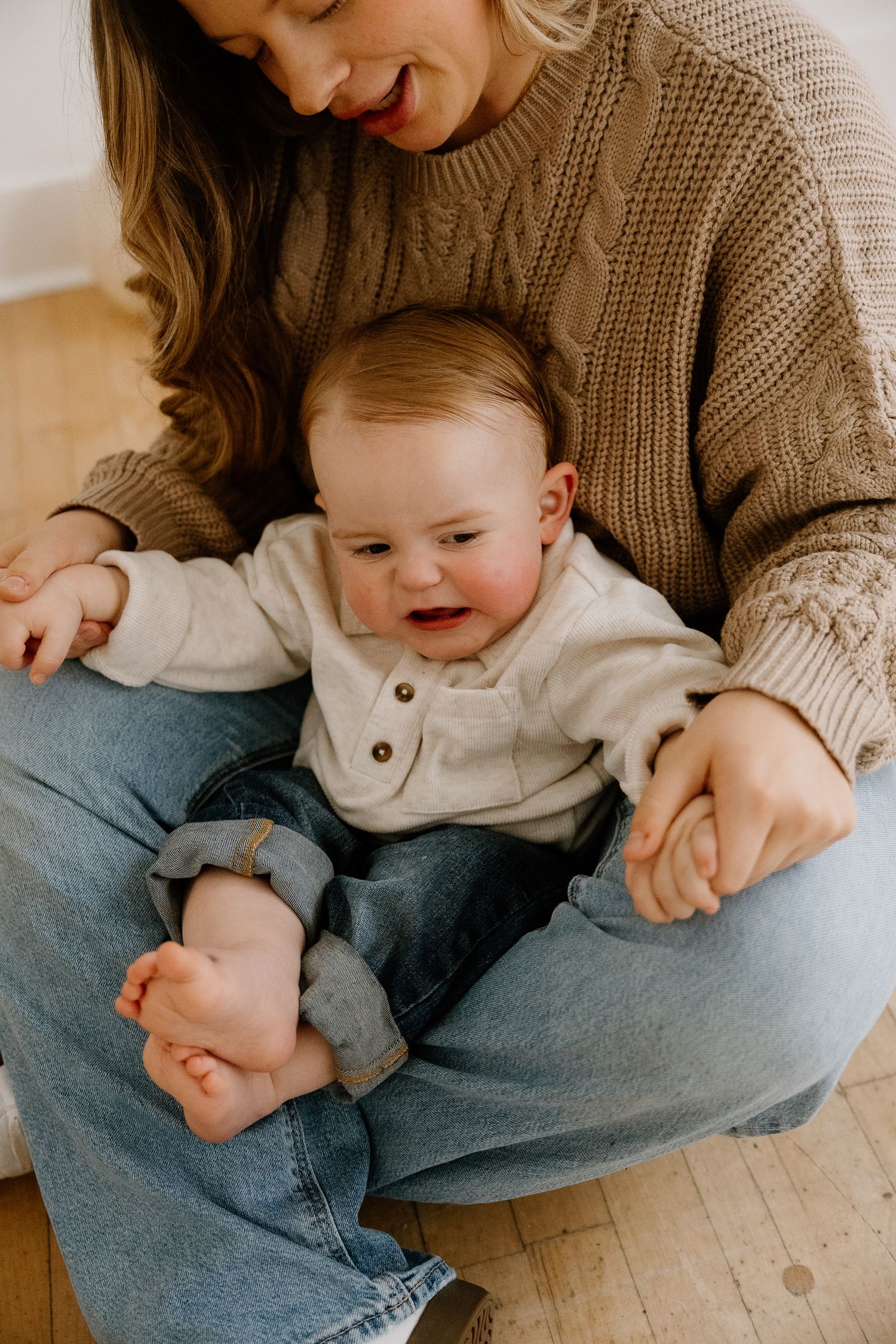 Mother in tan knit sweater sits cross-legged on wood floor cradling curious barefoot baby.