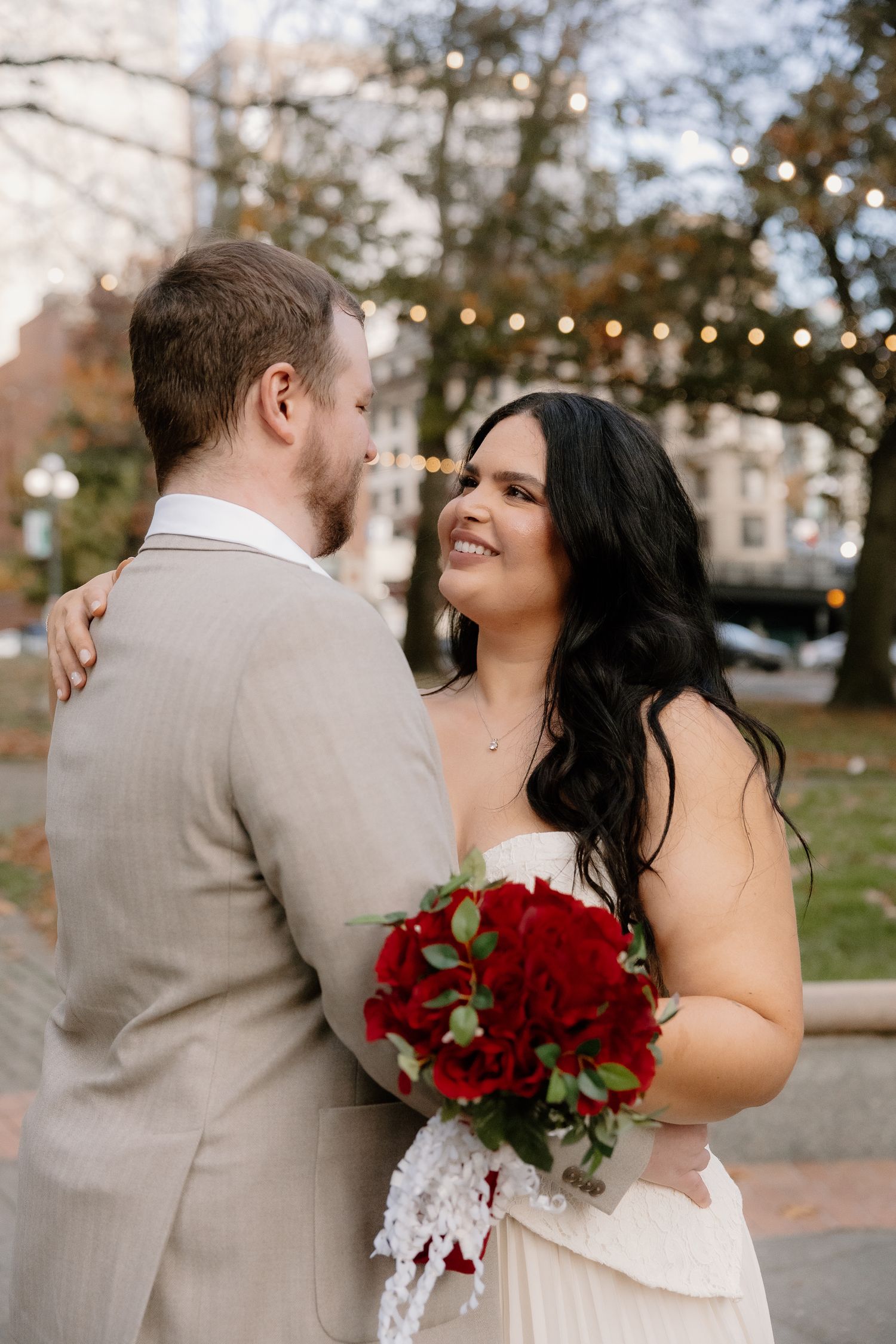 Smiling bride holds red rose bouquet while embracing groom outdoors with string lights behind.