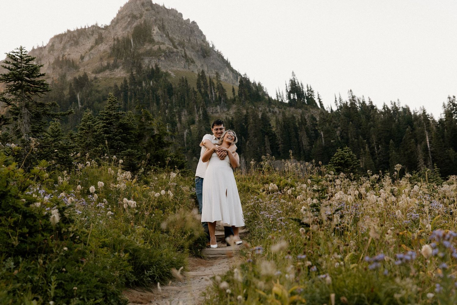 Couple embracing on a wildflower meadow trail with a dramatic mountain and forest backdrop.