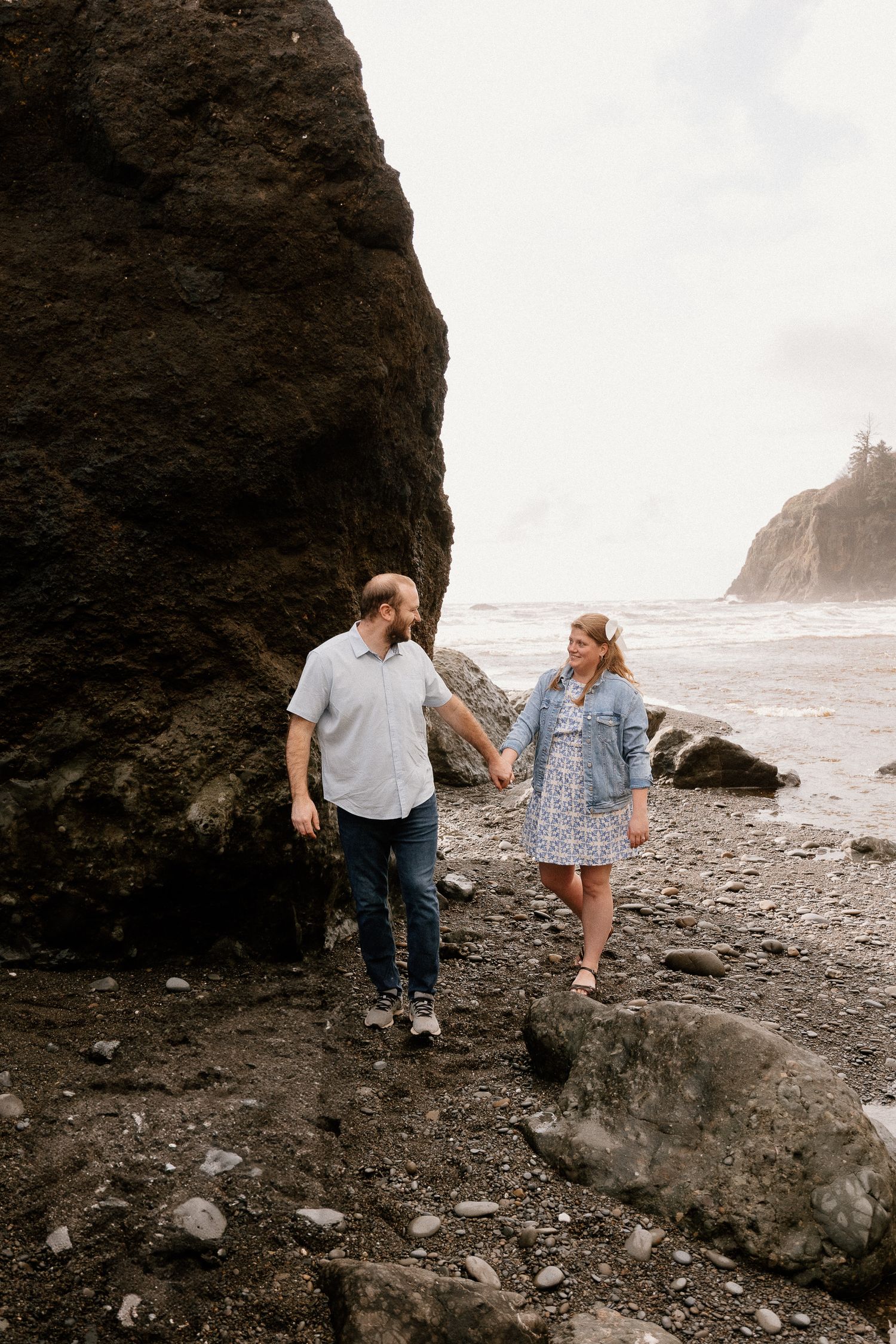 Couple holding hands strolling along rocky beach beside large dark sea stack with ocean waves behind.