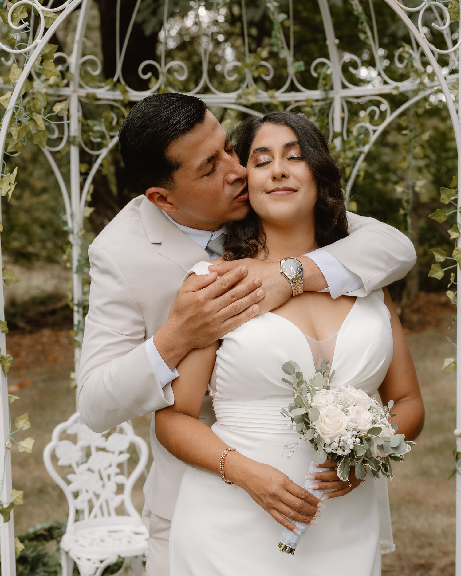 Groom in white suit kissing bride holding white floral bouquet under decorative garden arbor.