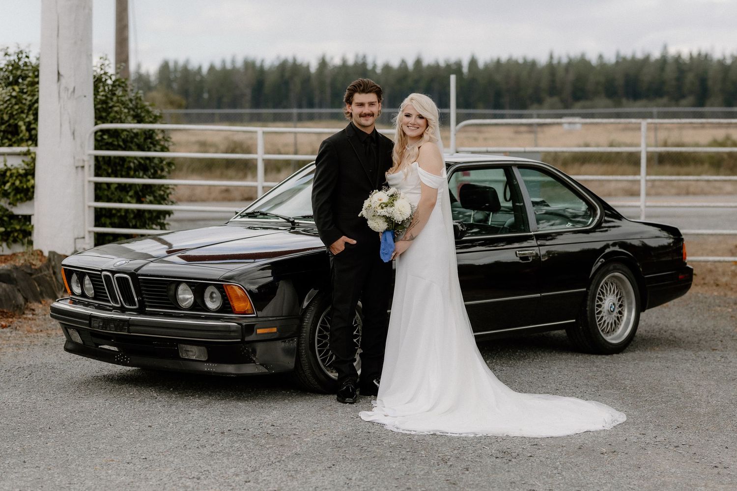 Bride and groom pose together in front of a classic black BMW at outdoor wedding venue.