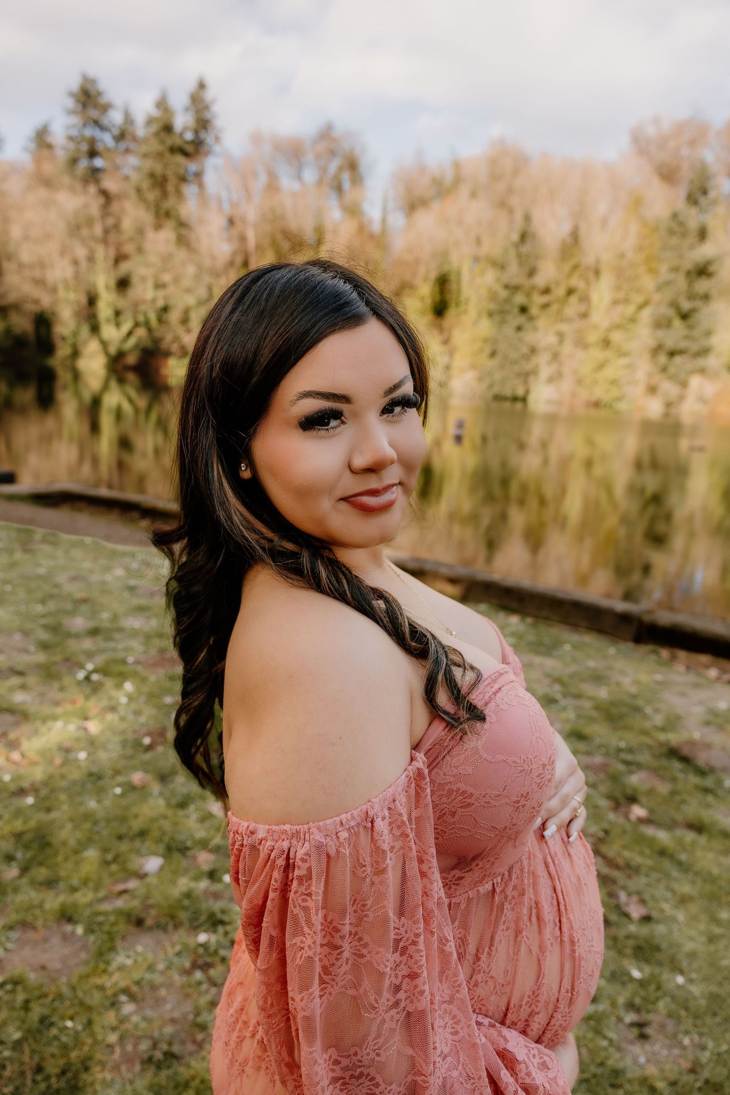 Pregnant woman in pink off-shoulder lace dress posing by a lake surrounded by autumn trees.
