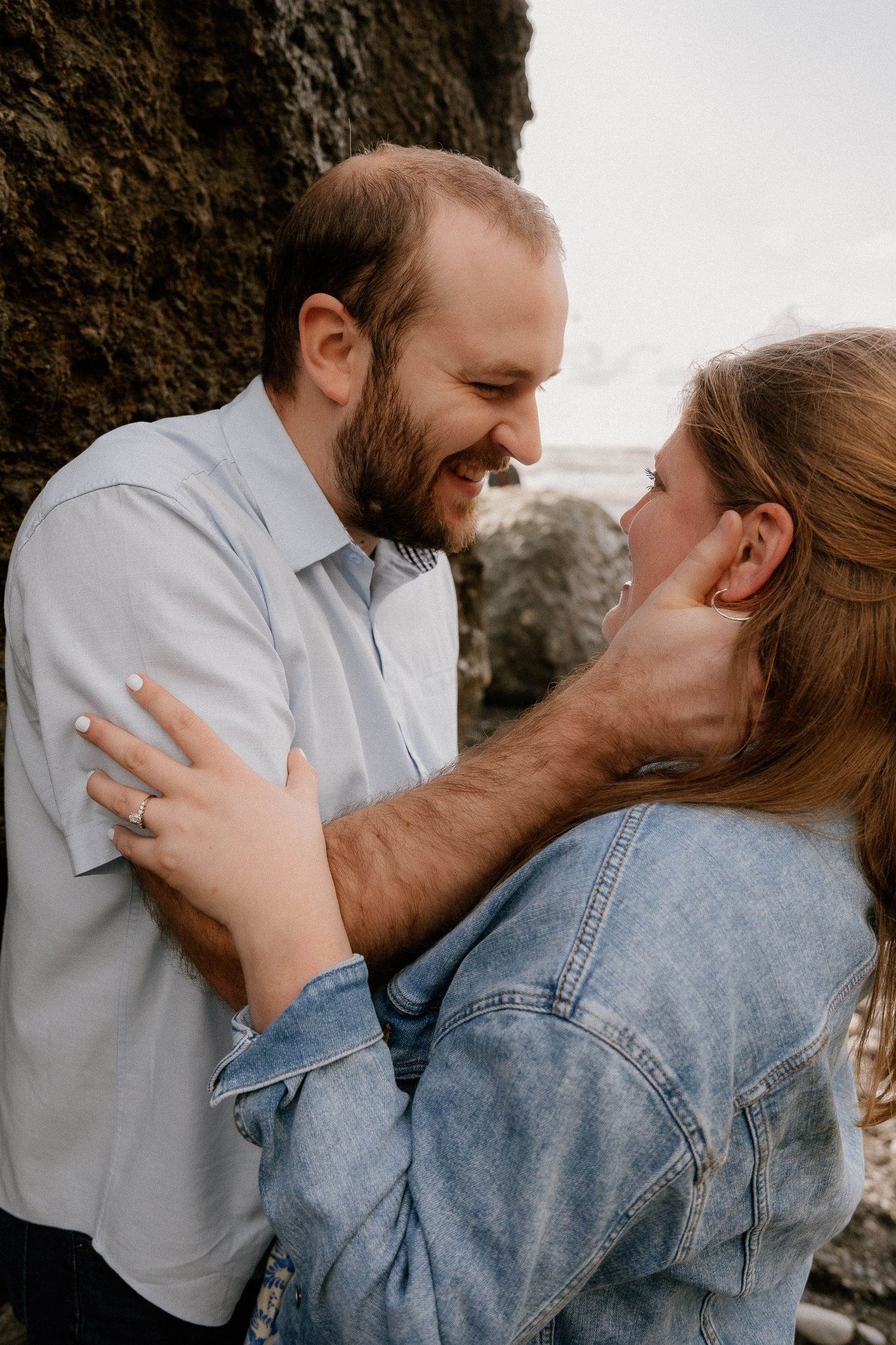 A smiling couple shares a romantic moment, the woman wearing a denim jacket and engagement ring, embracing near rocky cliffs.