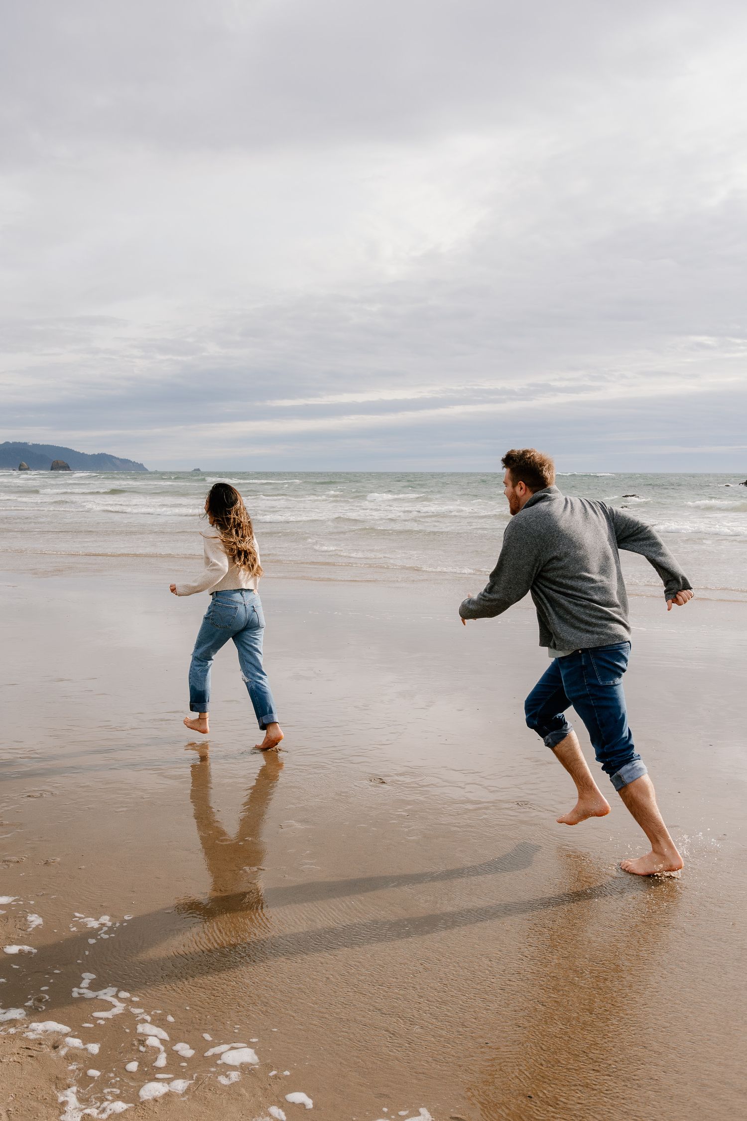 Couple playing barefoot on a sandy beach with ocean waves and cloudy sky in the background.