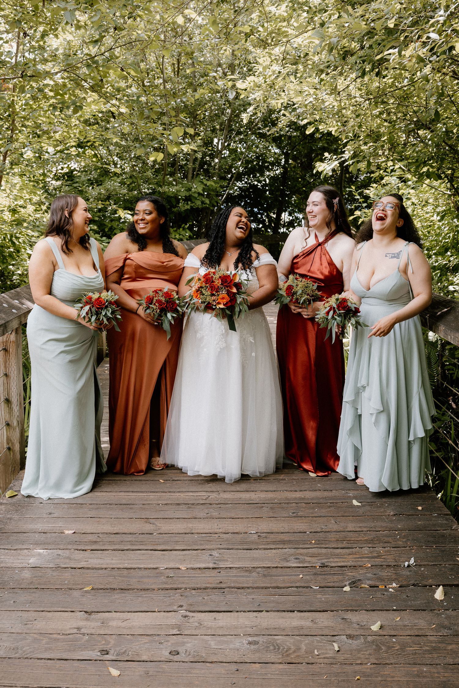 Bride laughing joyfully with four bridesmaids in mixed earth-tone gowns on wooden bridge outdoors.